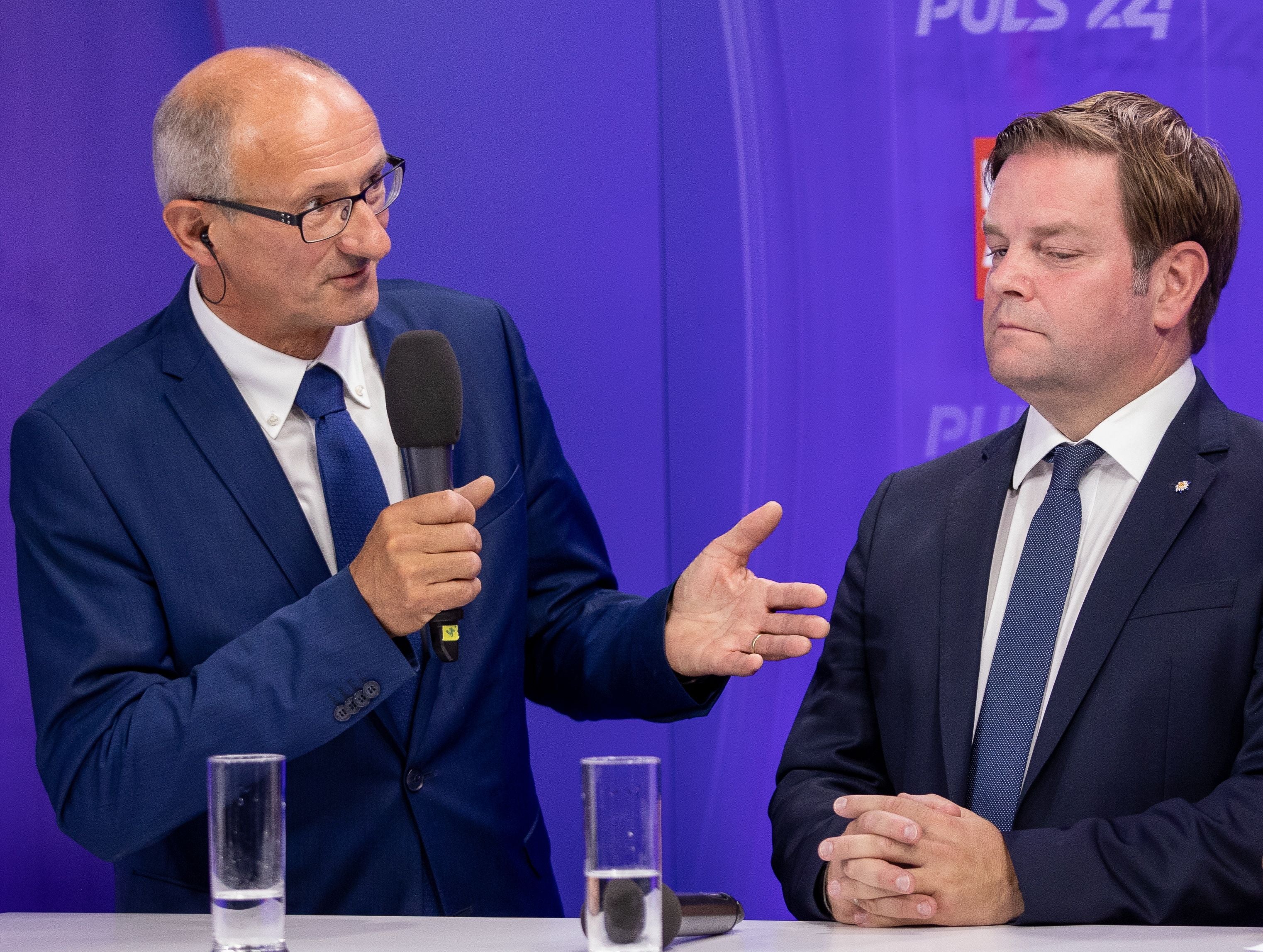 Zwei Männer in Anzügen bei einer Diskussion vor lila Hintergrund mit Mikrofon und Wassergläsern.
