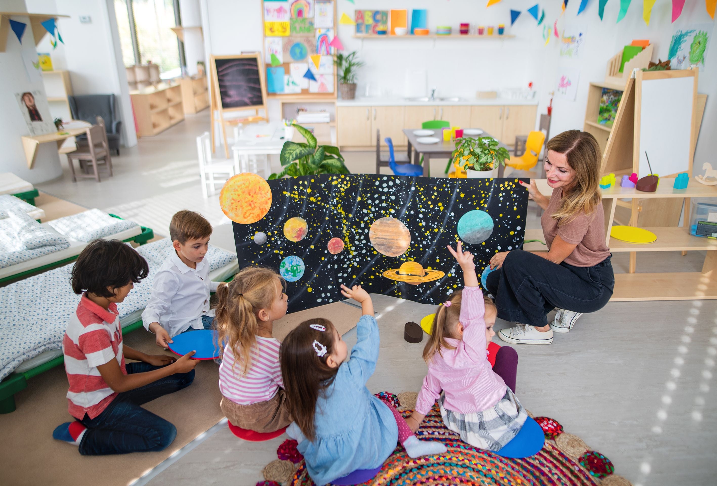 Lehrerin zeigt Kindern im Kindergarten ein Plakat mit Planeten und Sternen im Sonnensystem.