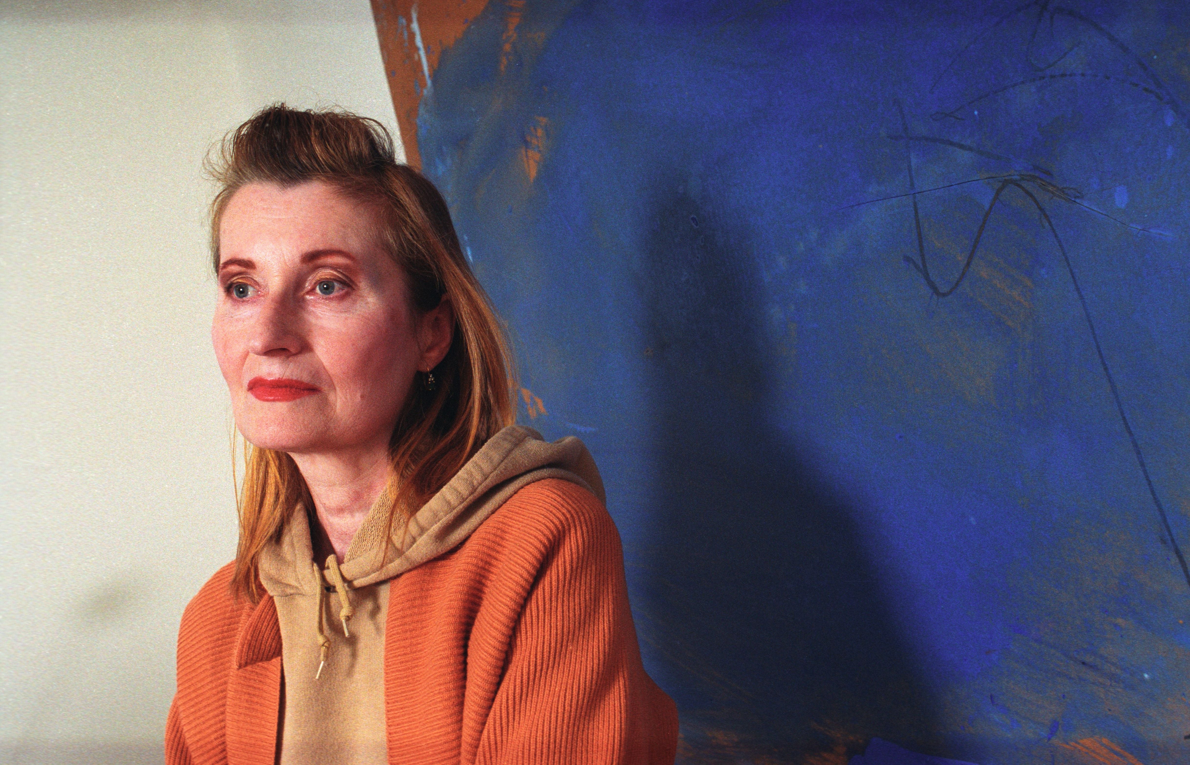 Frau mit orangefarbener Jacke vor blauem Hintergrund, blickt ernst nach links.