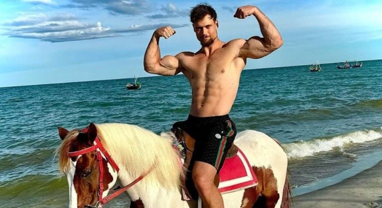 Ein muskulöser Mann posiert oberkörperfrei auf einem Pferd am Strand.