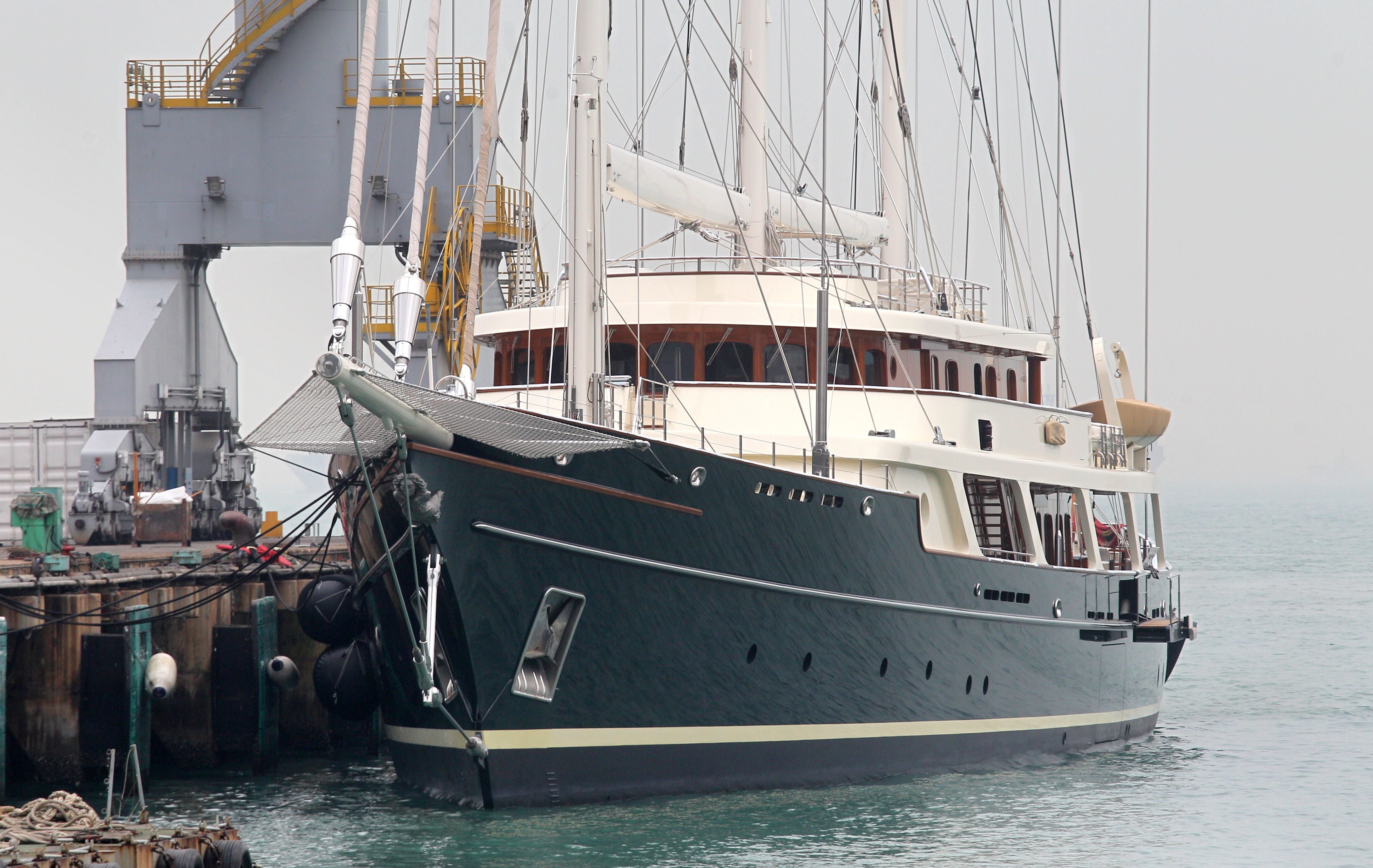 Dreimastige Segelyacht liegt im Hafen an einem Pier vor Industrieanlagen.