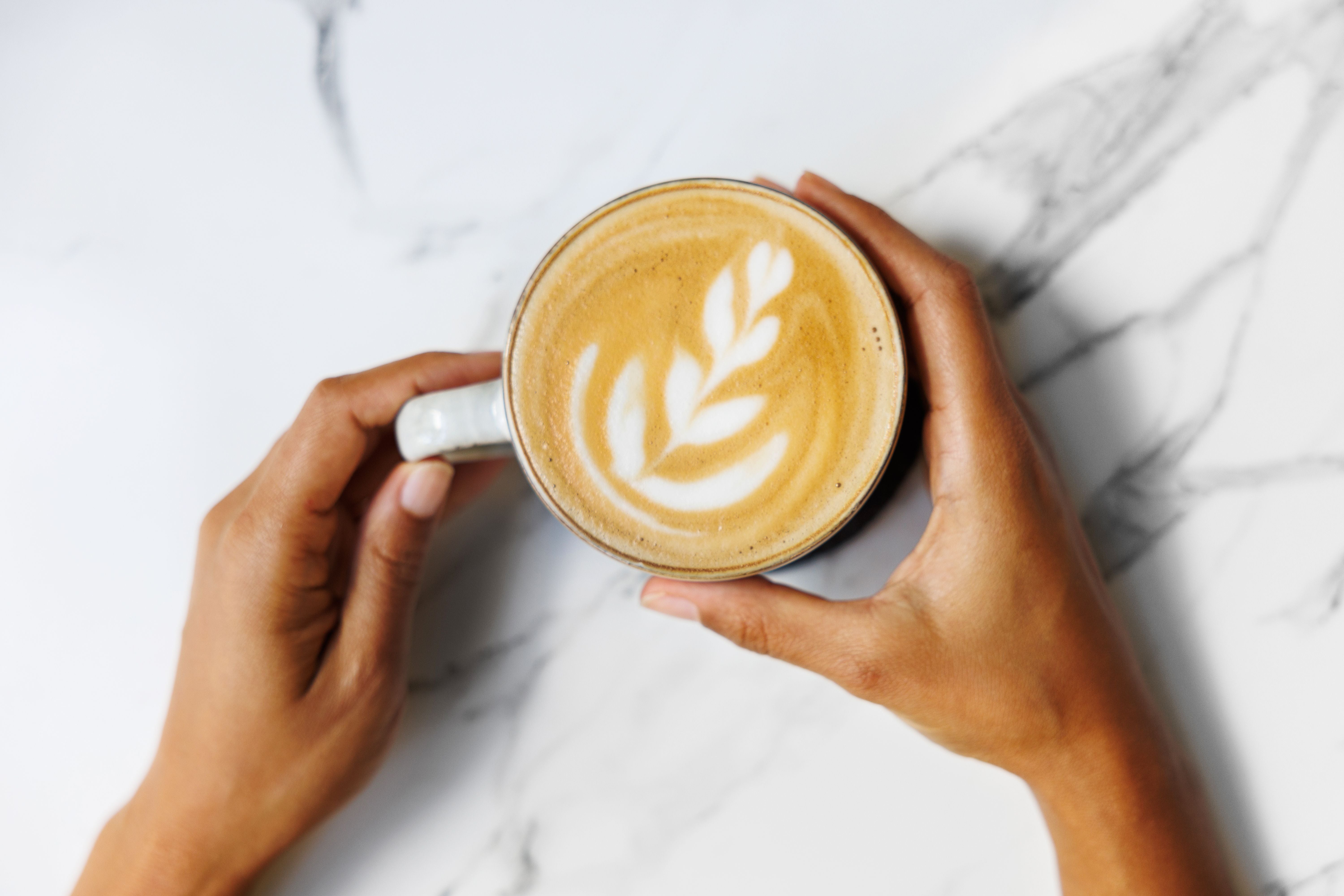 Hände halten eine Tasse Kaffee mit Latte Art auf einer weißen Marmorplatte.
