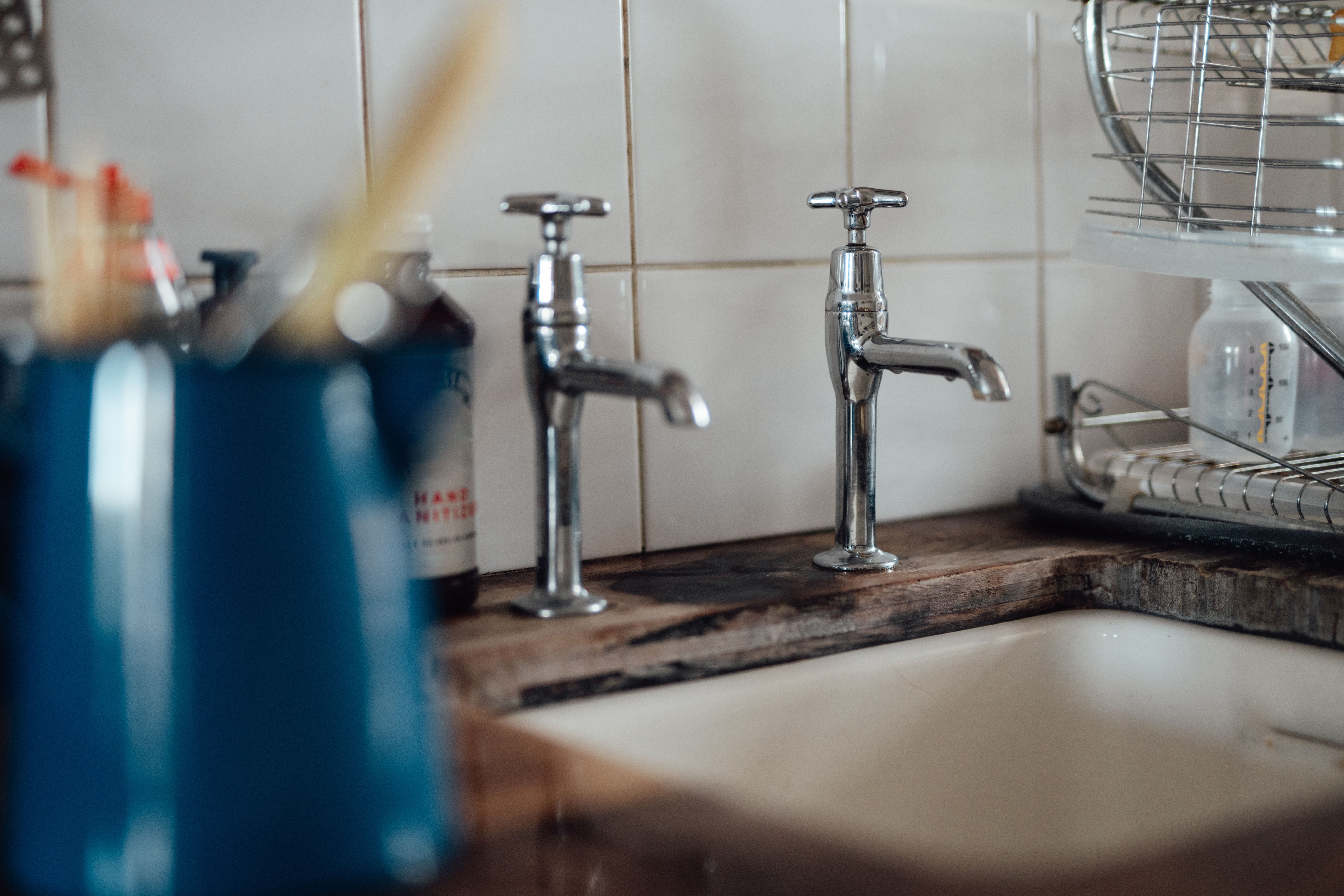 Zwei silberne Wasserhähne an einem Spülbecken mit weißem Fliesenspiegel im Hintergrund.