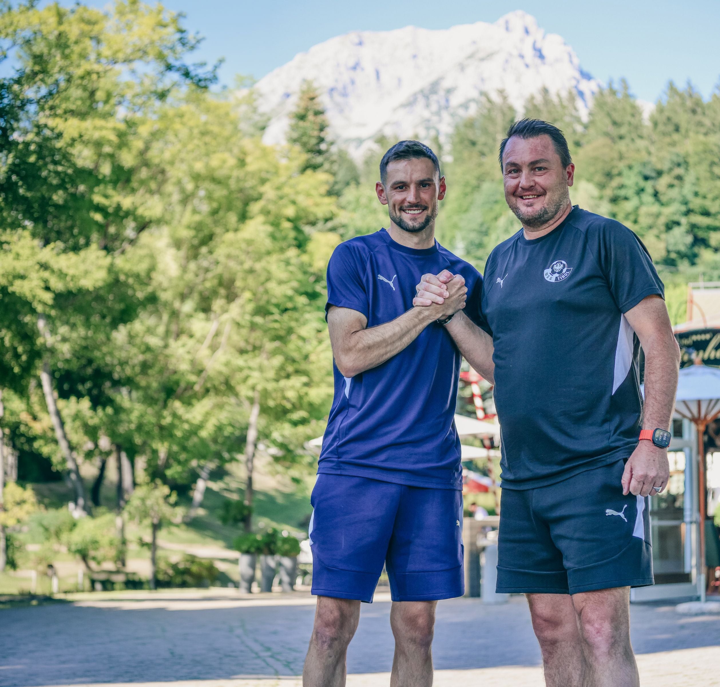 Zwei Männer im sportlichen Trainingsgewand stehen im Freien vor Bäumen und einem bergigen Hintergrund. Sie lächeln in die Kamera und geben sich kräftig die Hand.