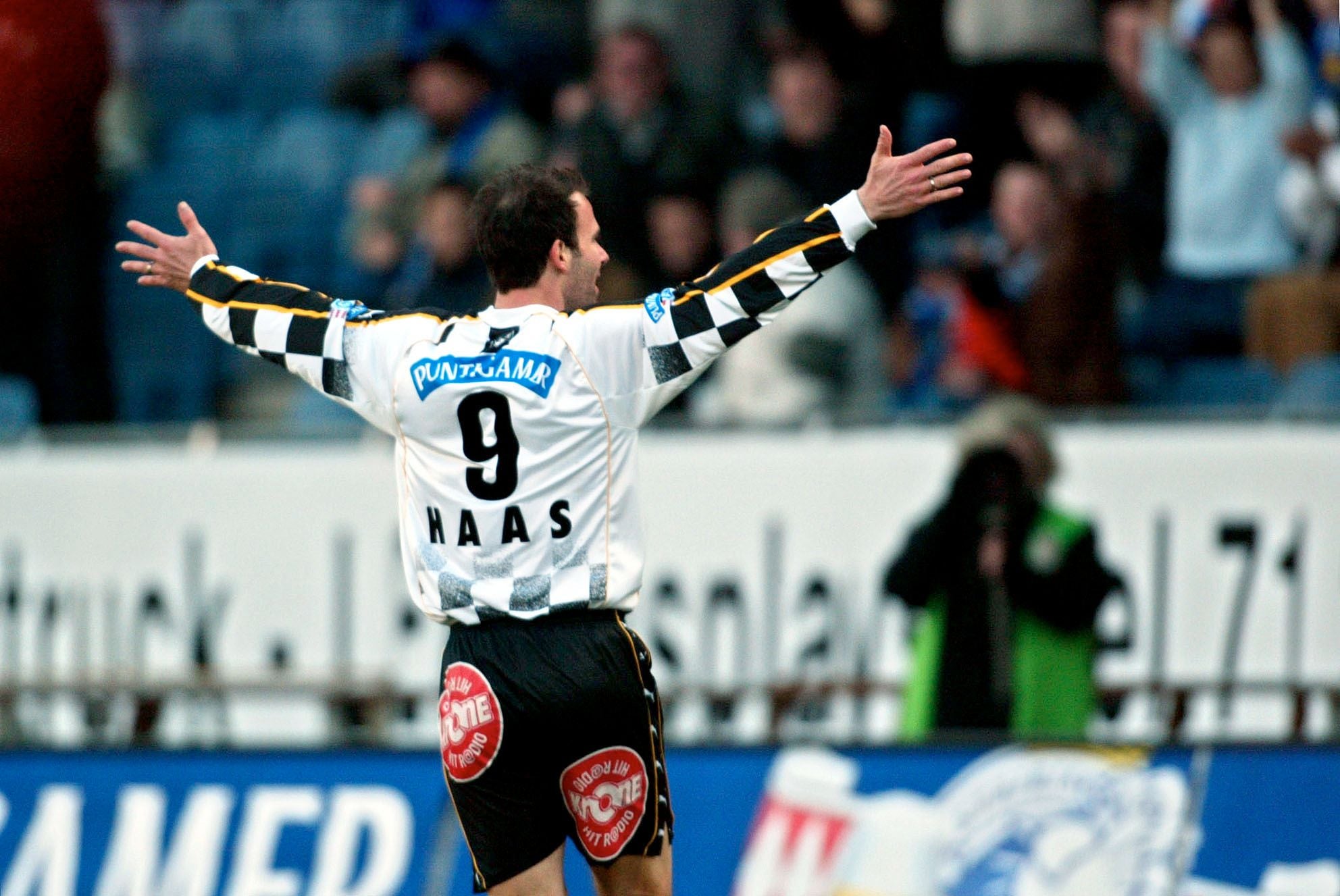 Ein Fußballspieler von hinten in einem weiß-schwarz karierten Trikot mit der Rückennummer 9 und dem Namen Haas breitet jubelnd die Arme aus. Im Hintergrund sind unscharf Zuschauer auf einer Tribüne zu sehen.