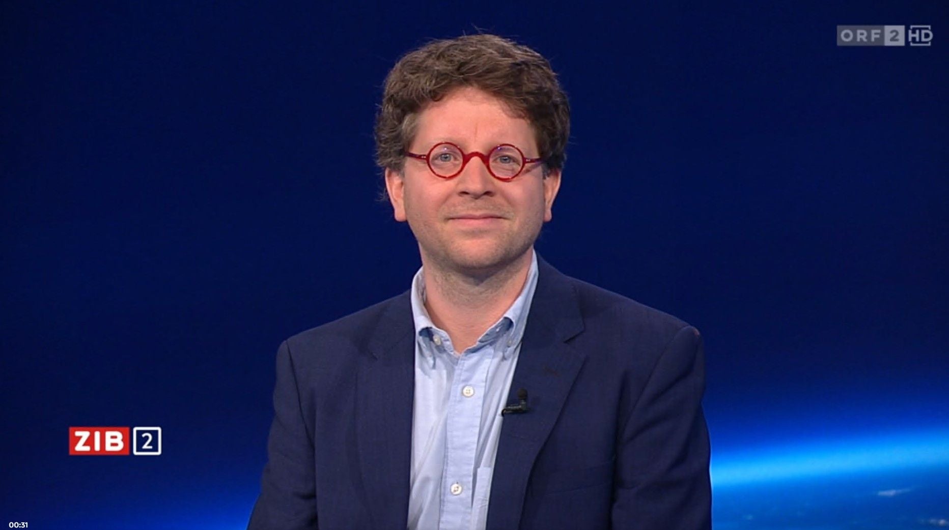 Ein Mann mit lockigen Haaren und roter Brille sitzt vor blauem Studiohintergrund.