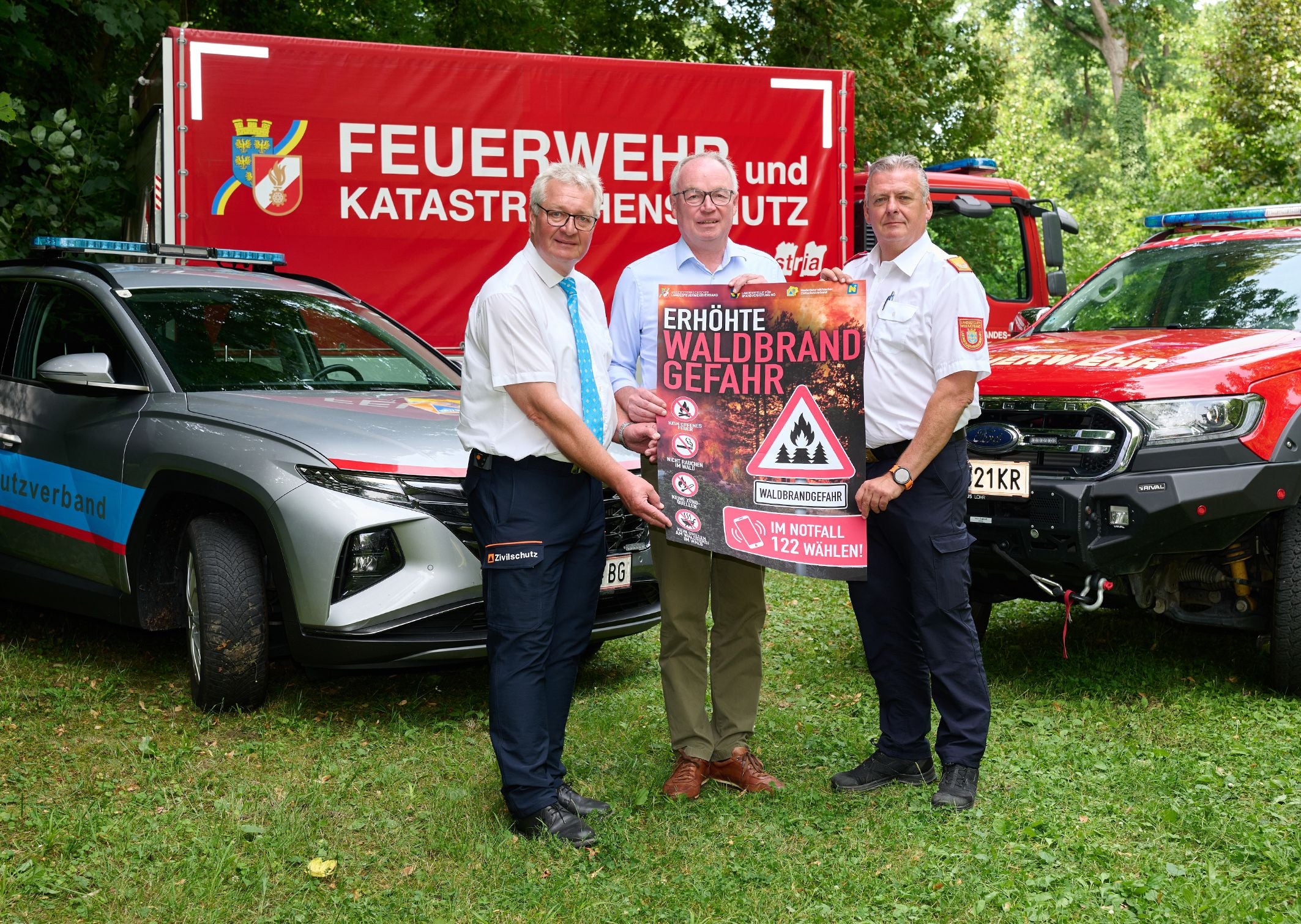 Drei Männer halten ein Schild zur erhöhten Waldbrandgefahr vor Feuerwehrfahrzeugen.