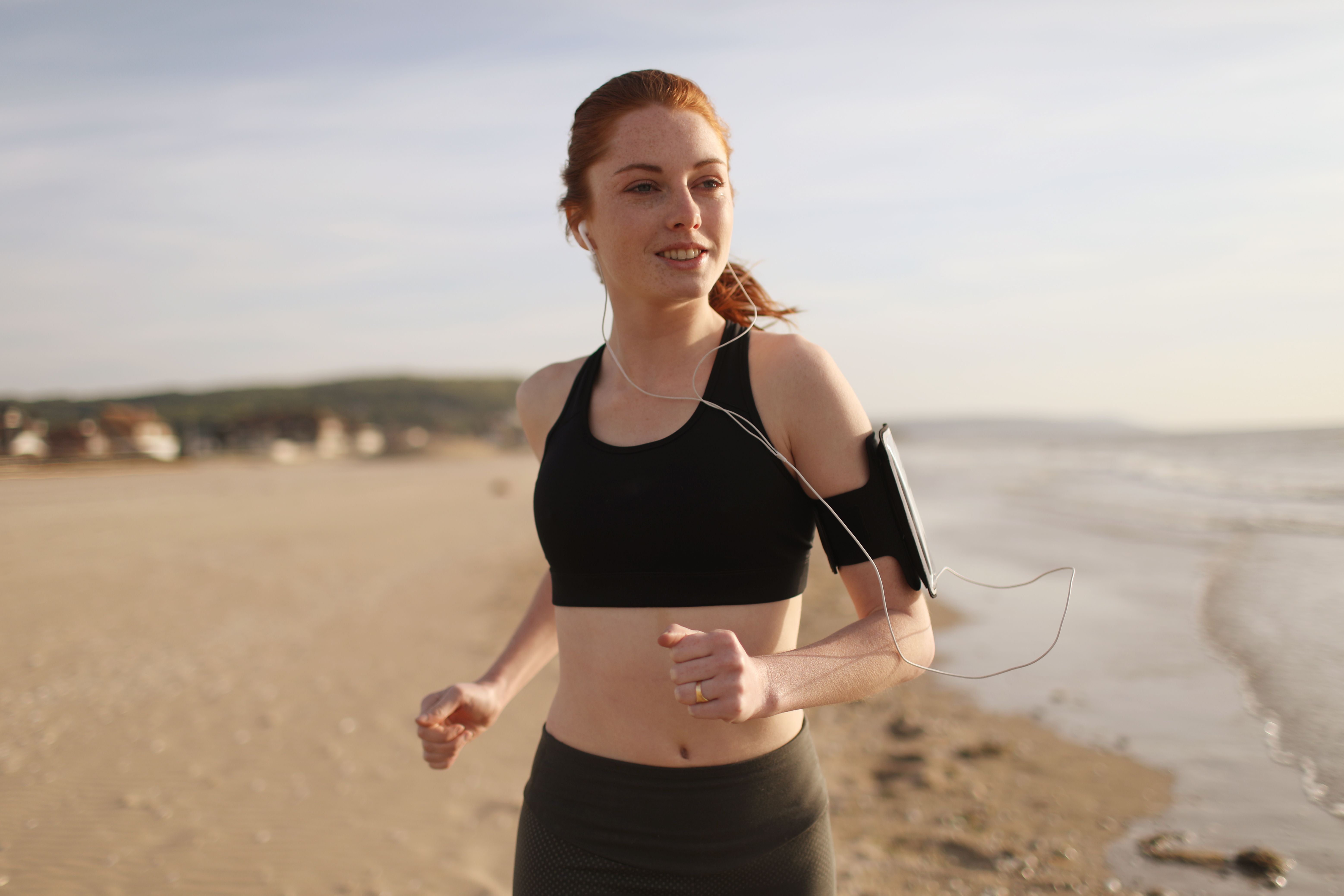 Junge Frau joggt am Strand mit Kopfhörern und Sportarmband bei sonnigem Wetter.