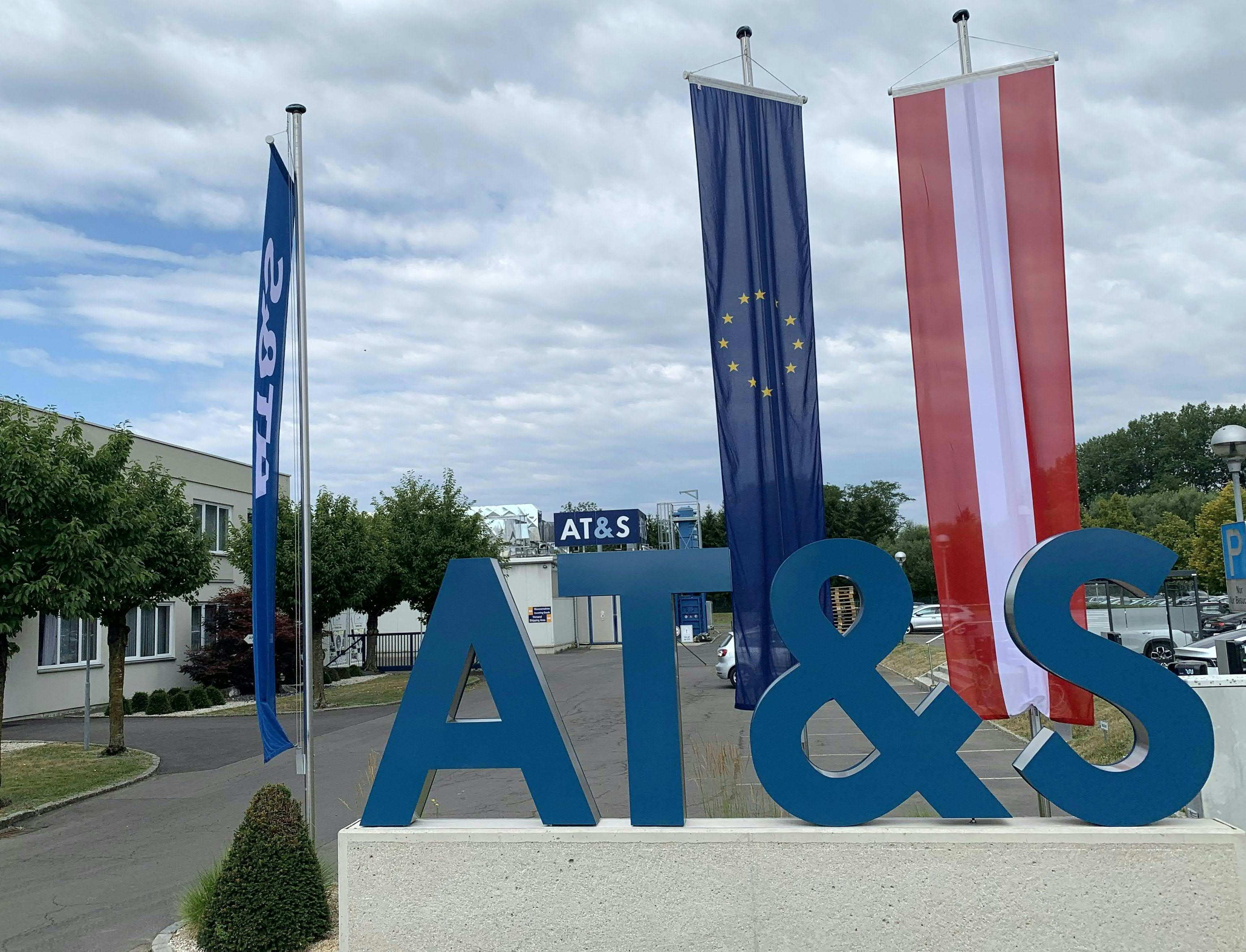 AT&S Logo vor Firmengebäude, daneben EU- und Österreich-Flaggen, sommerlicher Himmel.