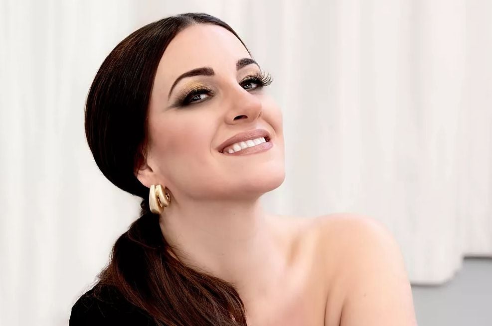 Frau mit braunen Haaren, Goldohrringen und elegantem Make-up lächelt vor hellem Hintergrund.