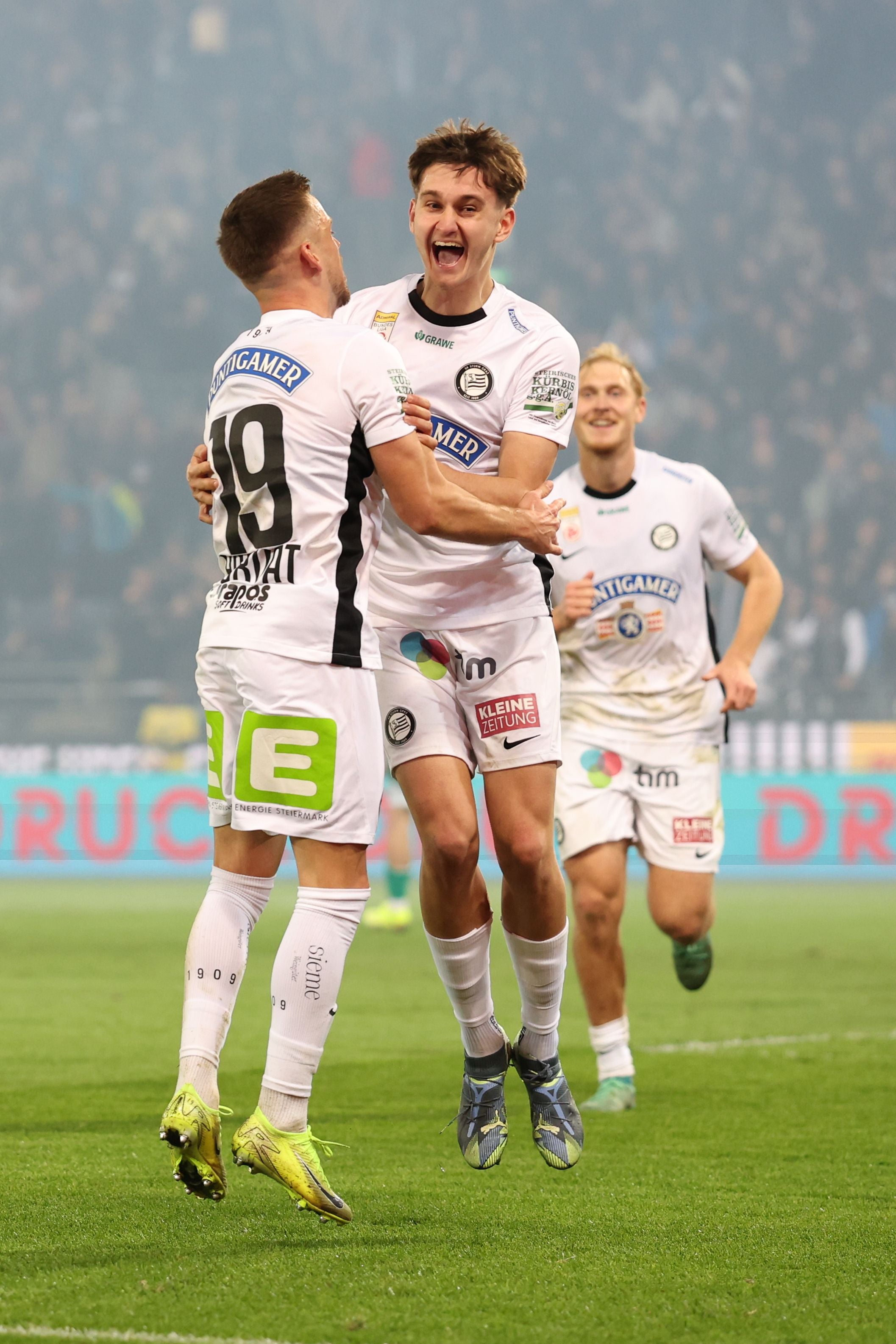 Zwei Fußballer im weißen Sturm-Graz-Trikot springen jubelnd in die Luft und umarmen sich auf dem Rasen. Ein dritter Spieler läuft lächelnd im Hintergrund auf sie zu.