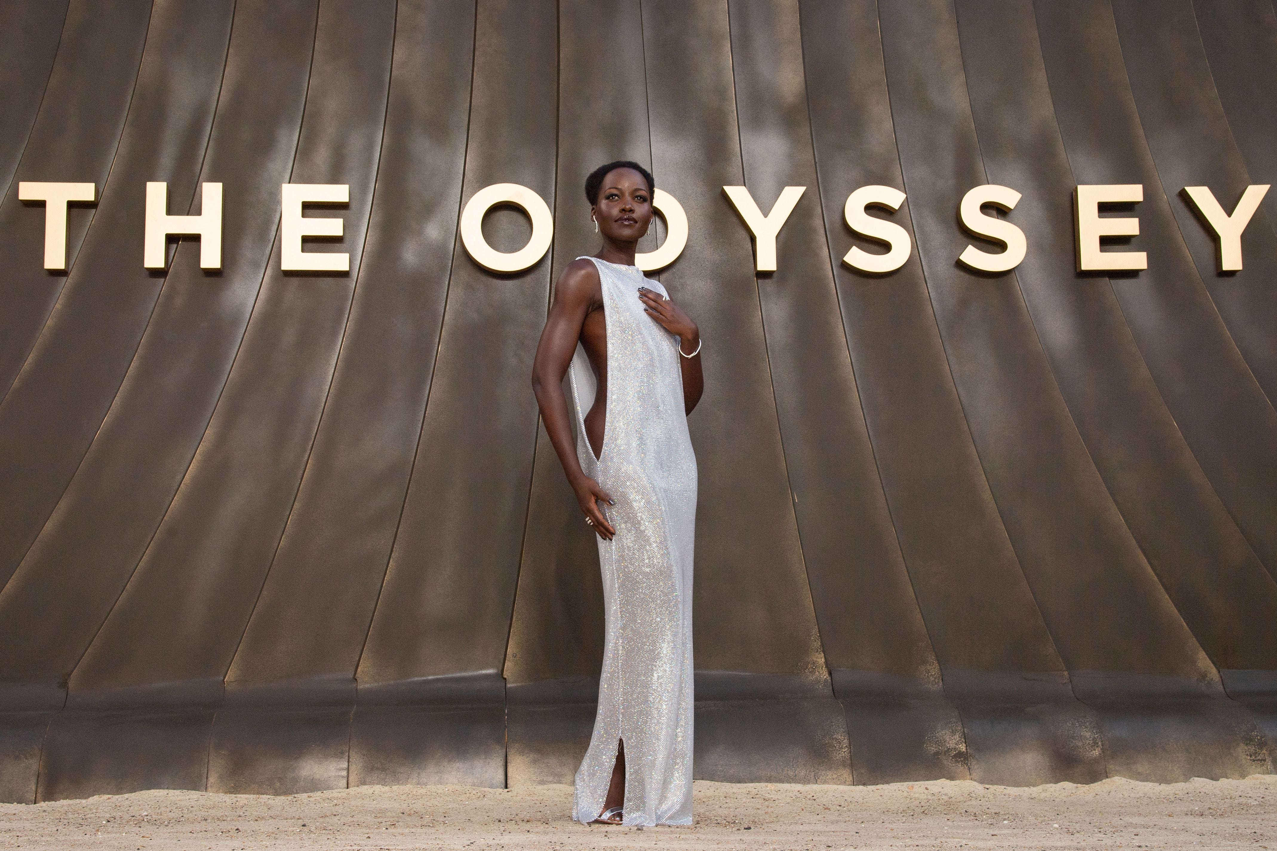 Eine Frau im silbernen Kleid posiert vor einer Wand mit dem Schriftzug „THE ODYSSEY“.