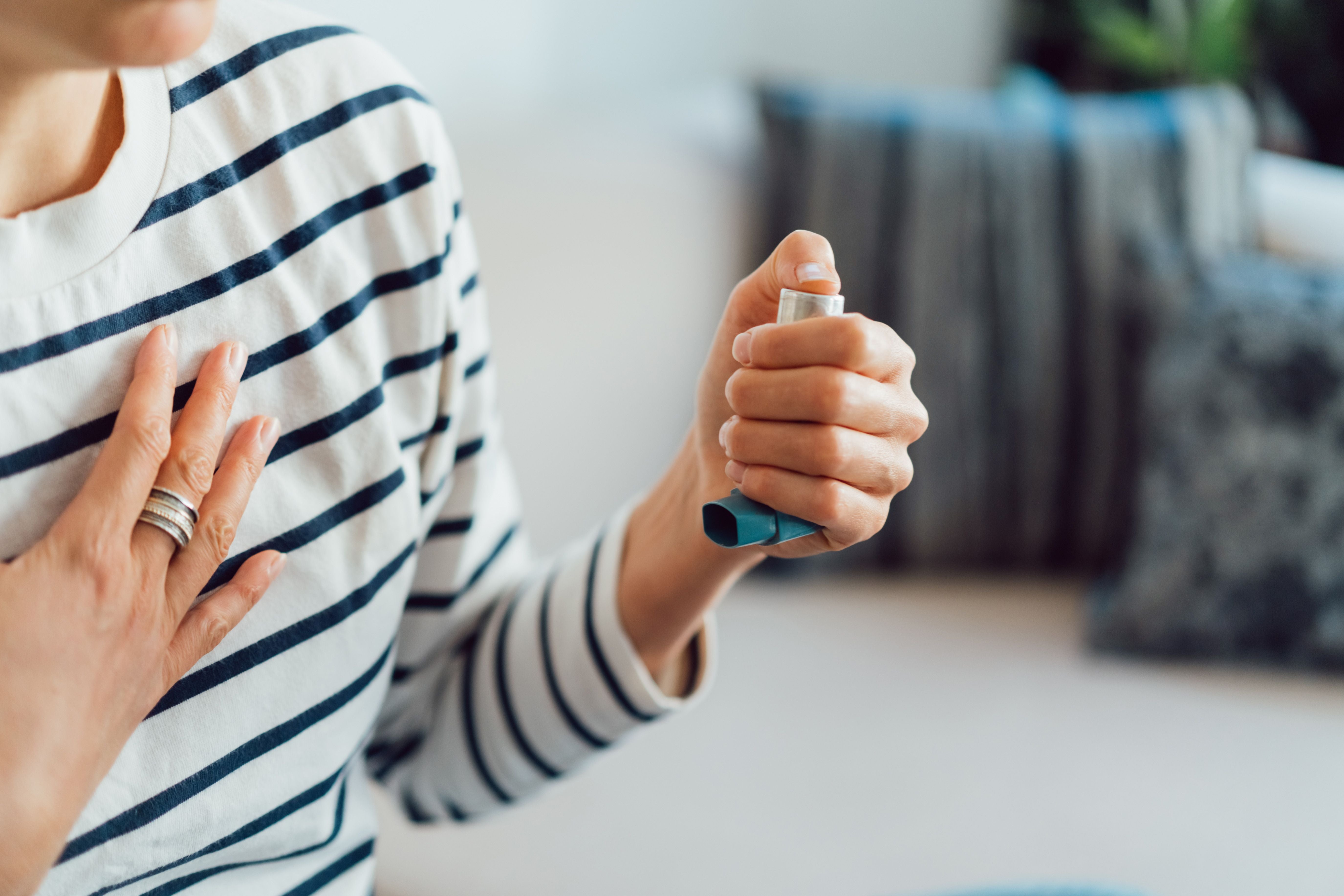 Frau hält einen Asthmainhalator und fasst sich an die Brust, vermutlich bei einem Asthmaanfall.