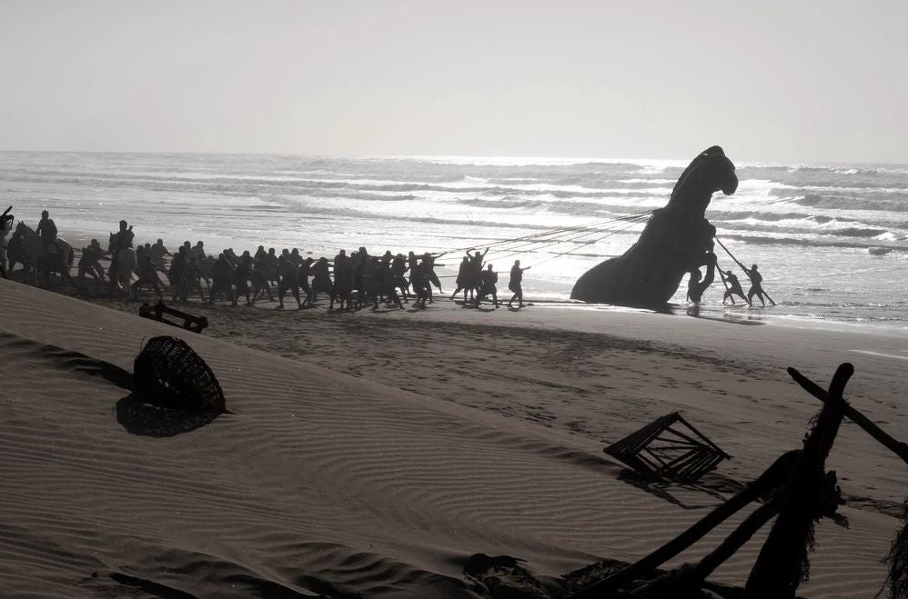 Viele Menschen ziehen ein großes hölzernes Pferd am Strand entlang zum Meer.