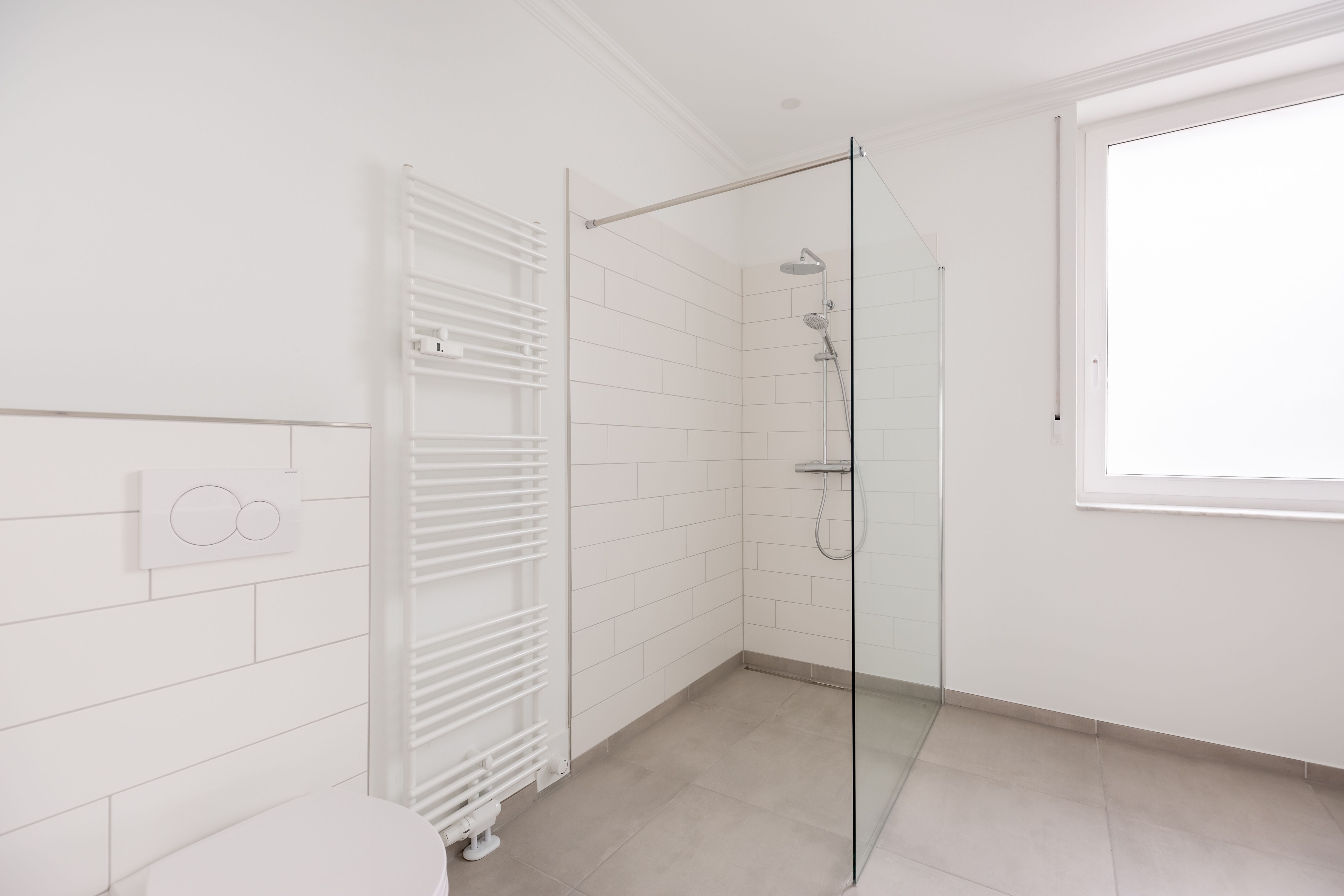 Modernes, weißes Bad mit bodengleicher Dusche, Handtuchheizkörper und großem Fenster.