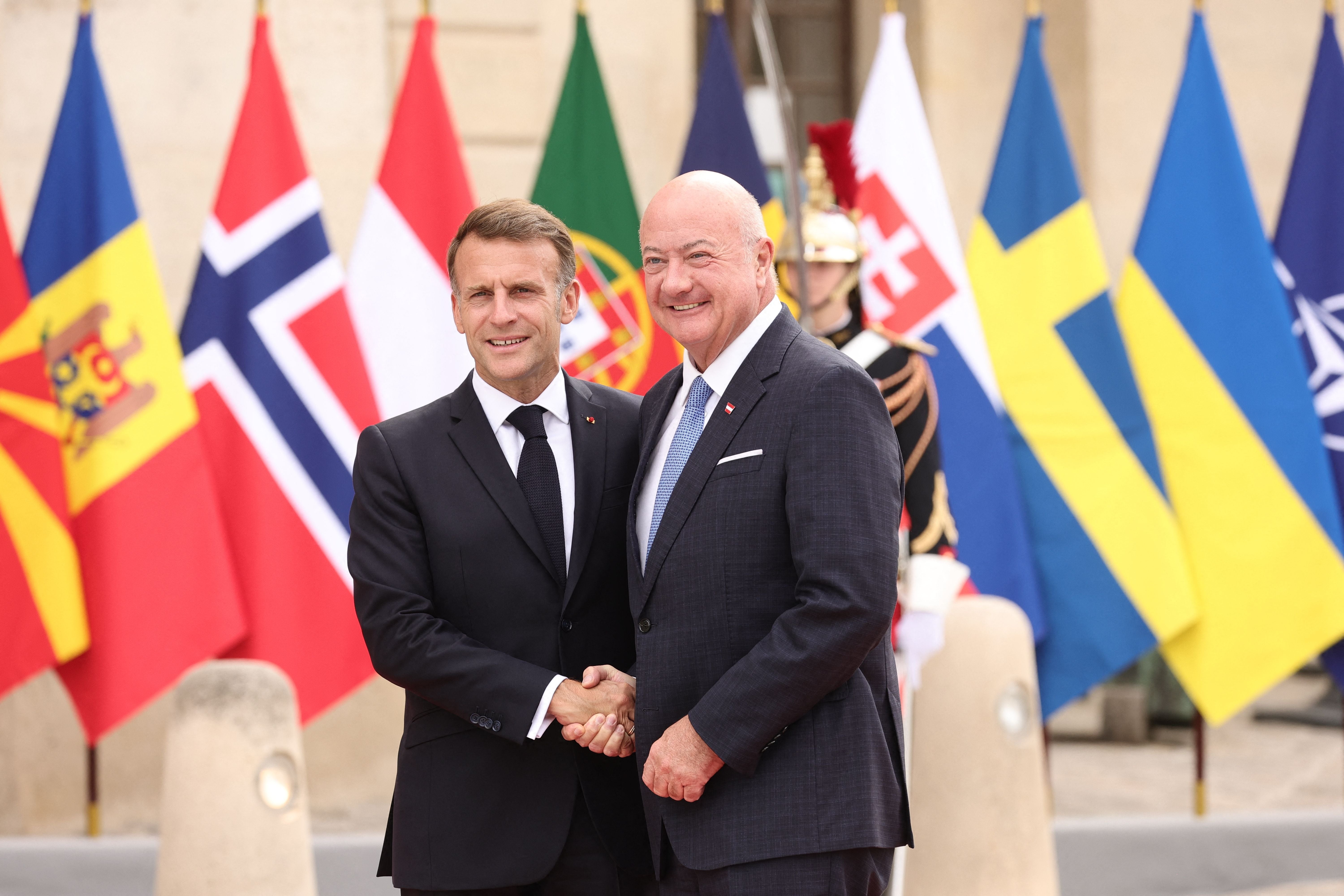 Emmanuel Macron und Christian Stocker schütteln sich vor internationalen Flaggen die Hand.