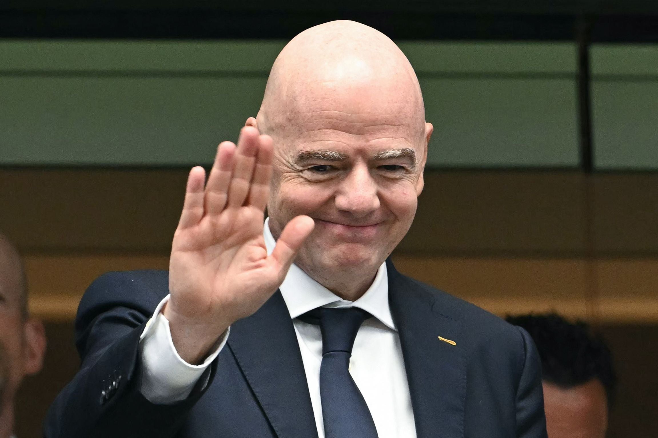 Ein Mann im Anzug winkt und lächelt in einer Zuschauerloge.