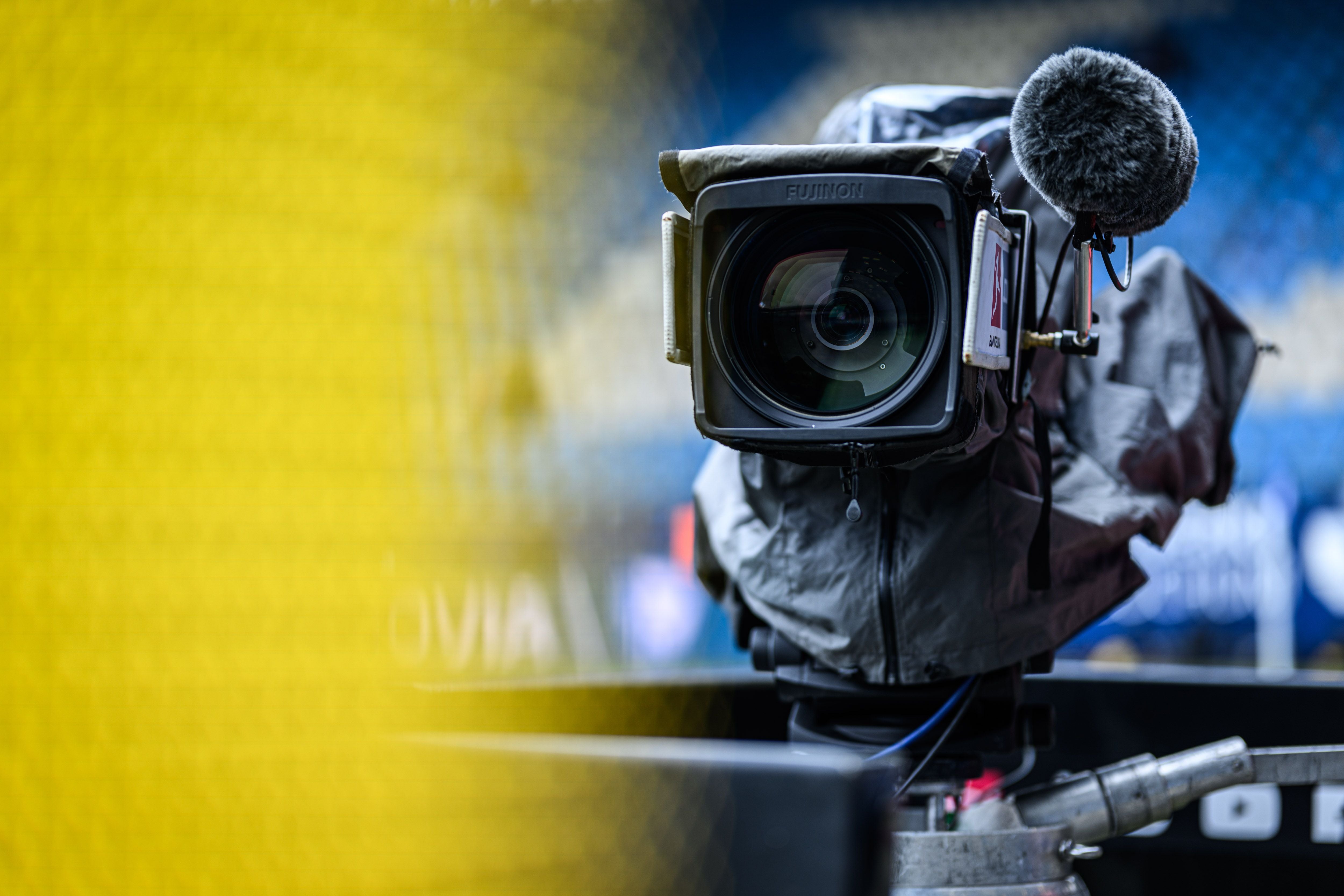 Fernsehkamera mit Mikrofon im Stadion, im Hintergrund unscharfe Sitzplätze und gelbes Netz.