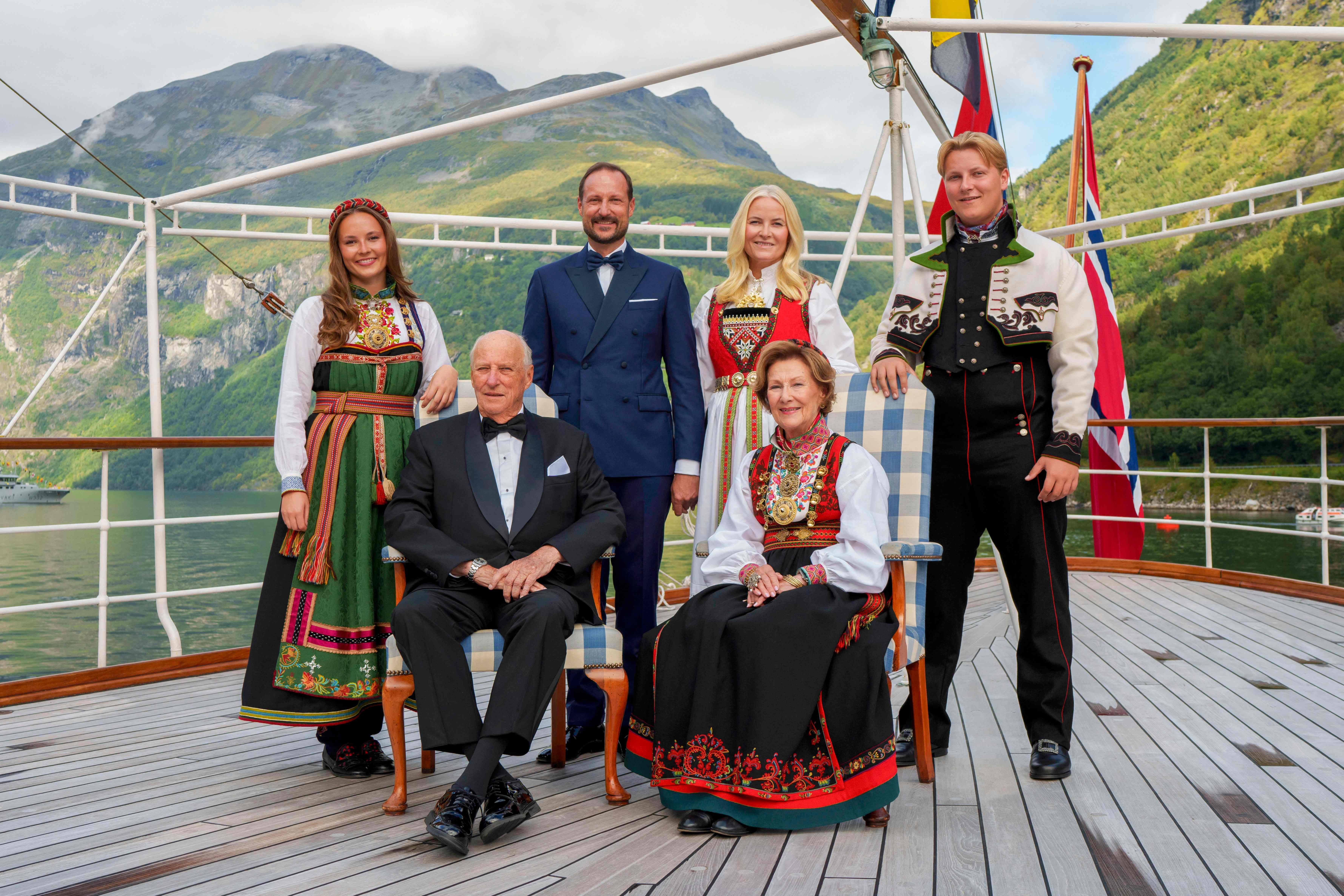 Sechs Personen in festlicher Tracht und Anzug posieren auf einem Boot vor norwegischer Fjordlandschaft.
