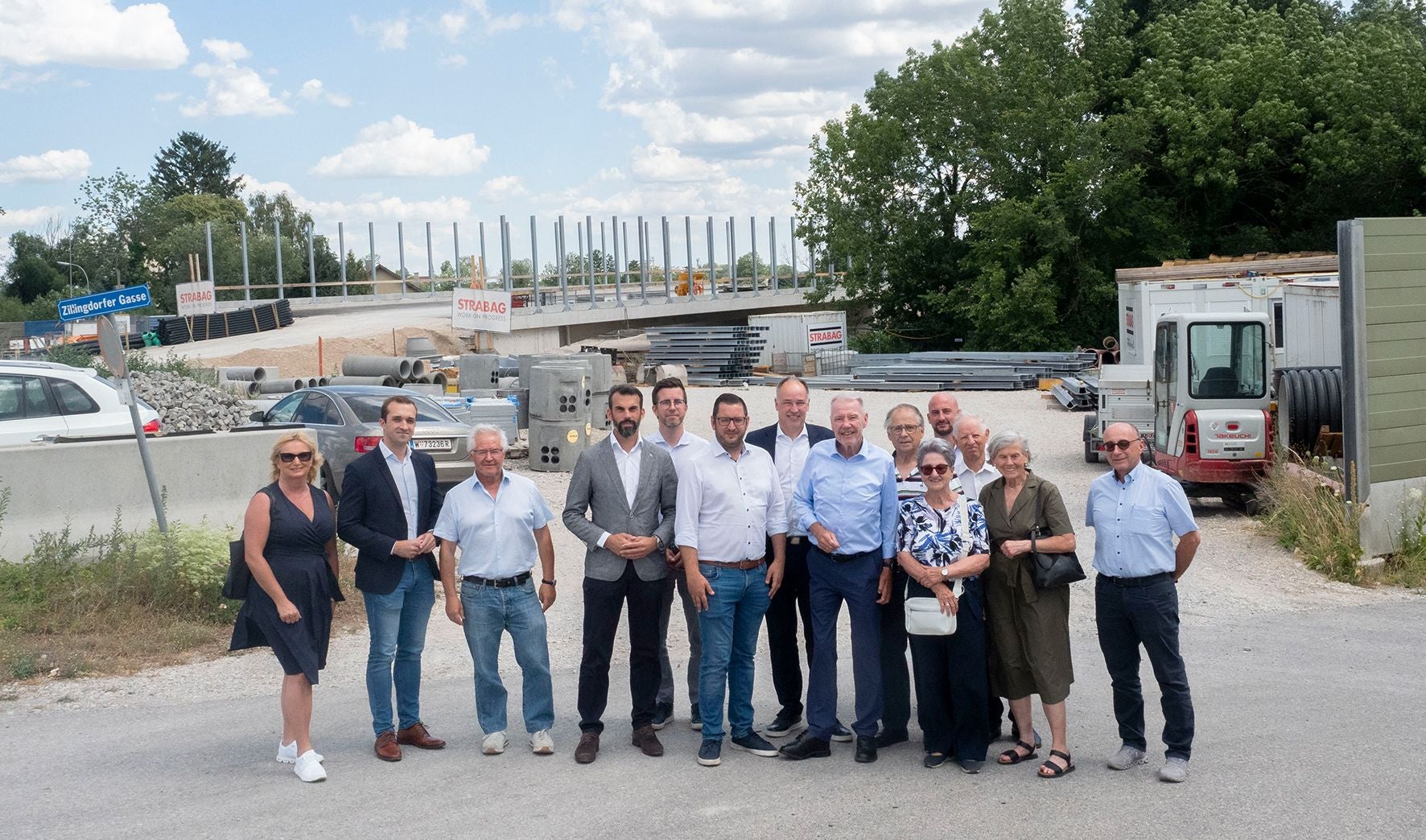 Eine Gruppe von Menschen steht vor einer Baustelle mit Brücke und Baugeräten im Hintergrund.