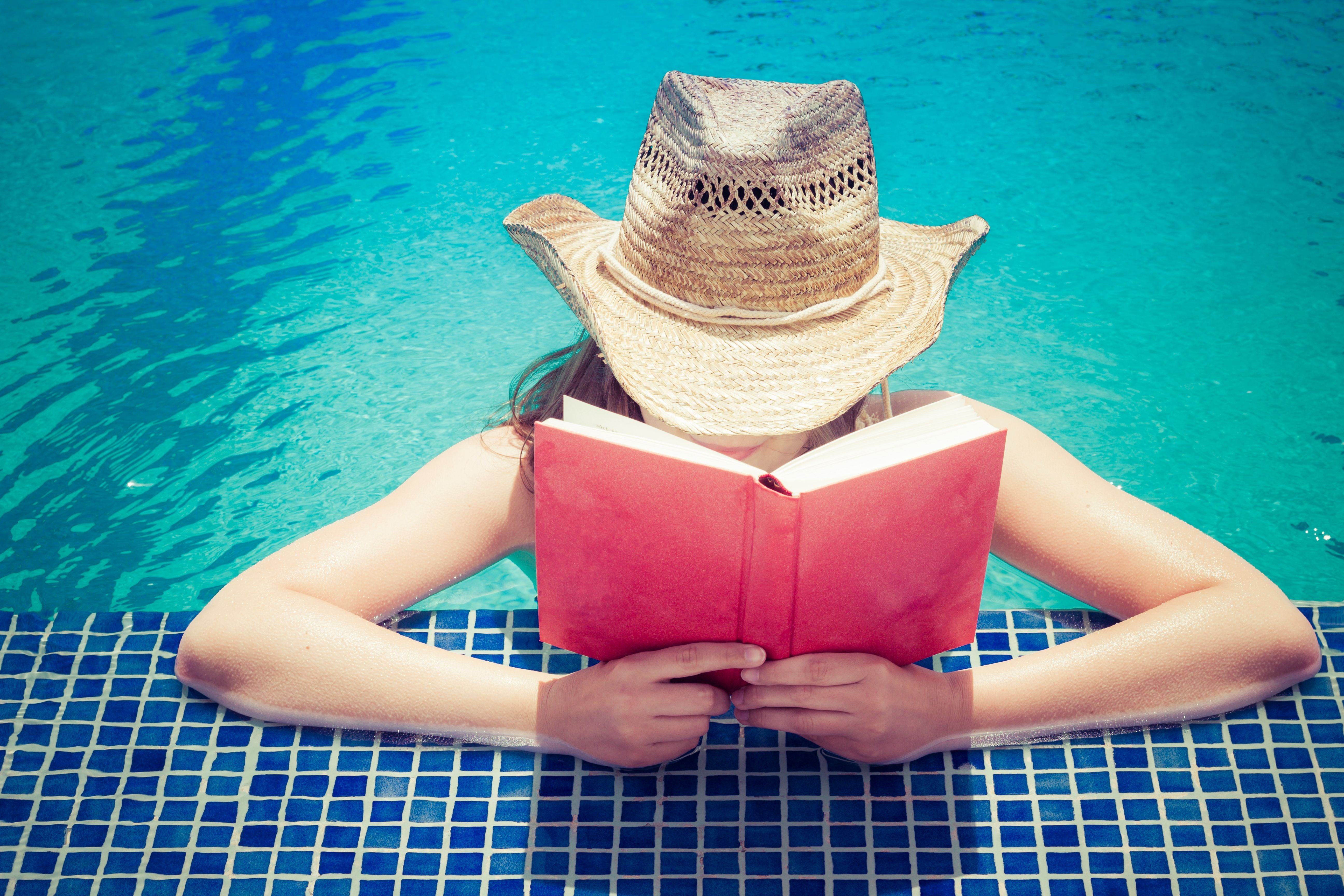 Person mit Strohhut liest ein Buch am Rand eines Pools im Sommer.