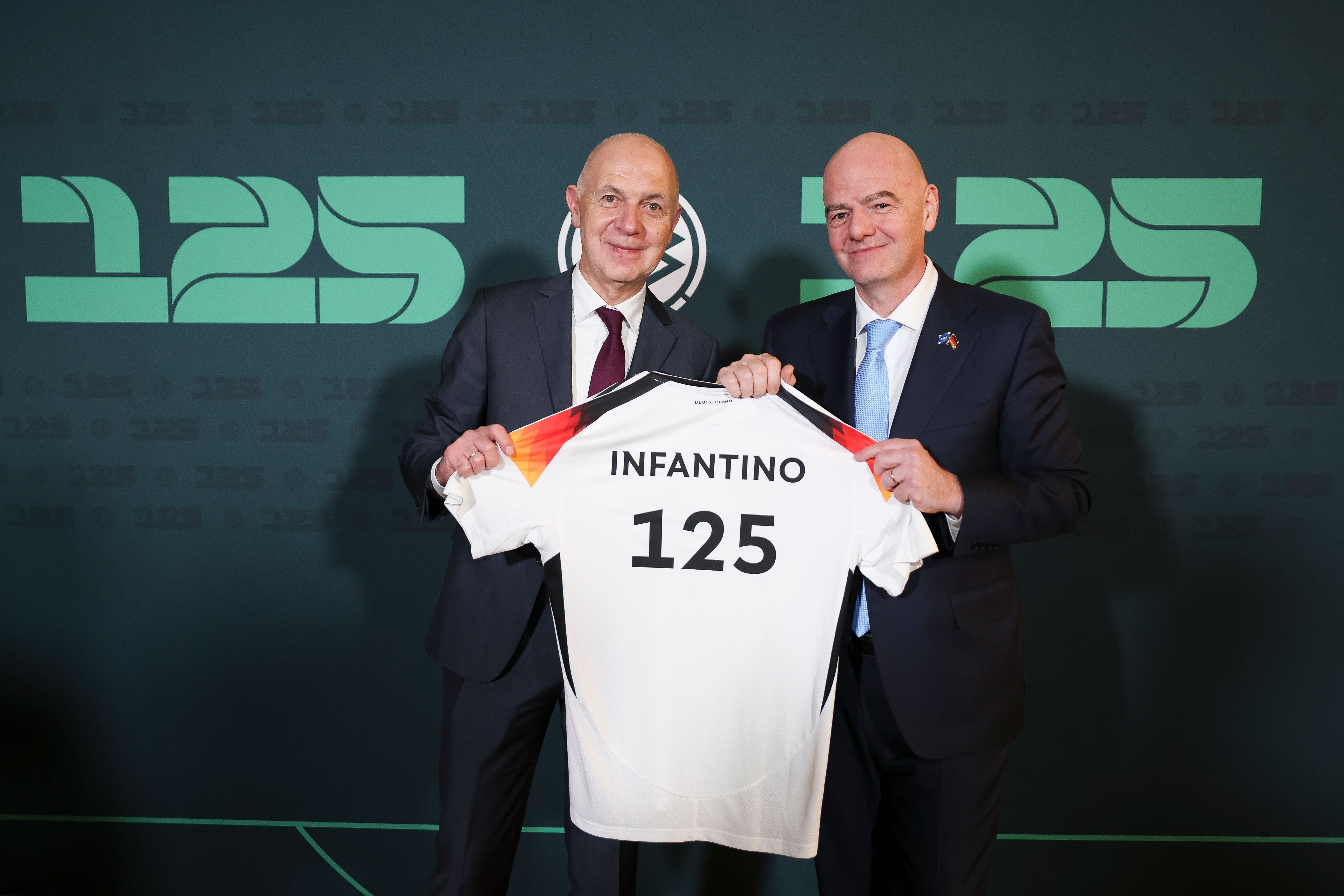 Zwei Männer im Anzug halten ein Trikot mit der Aufschrift "INFANTINO 125" vor grünem Hintergrund.