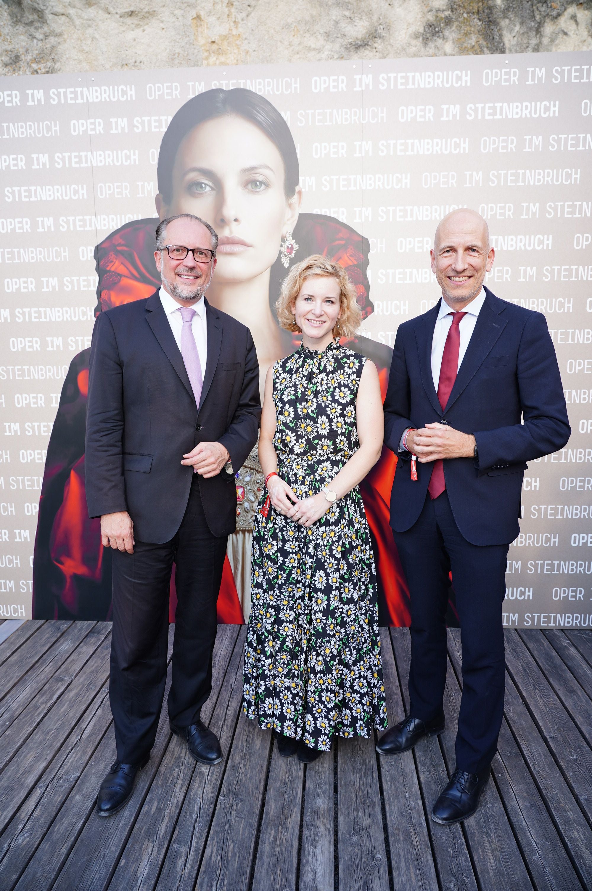 Drei Personen posieren vor einem "Oper im Steinbruch"-Tosca-Premiere-Plakat auf Holzplanken.