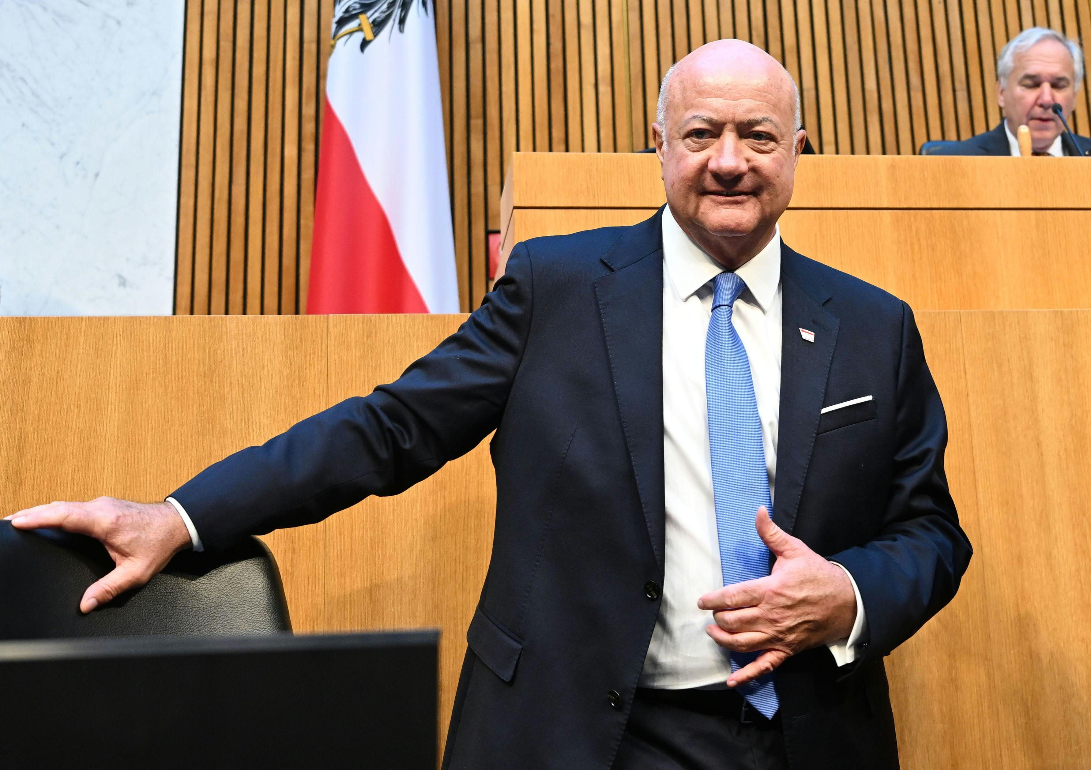 Ein Mann im Anzug steht im Parlament vor einer österreichischen Flagge und lächelt.