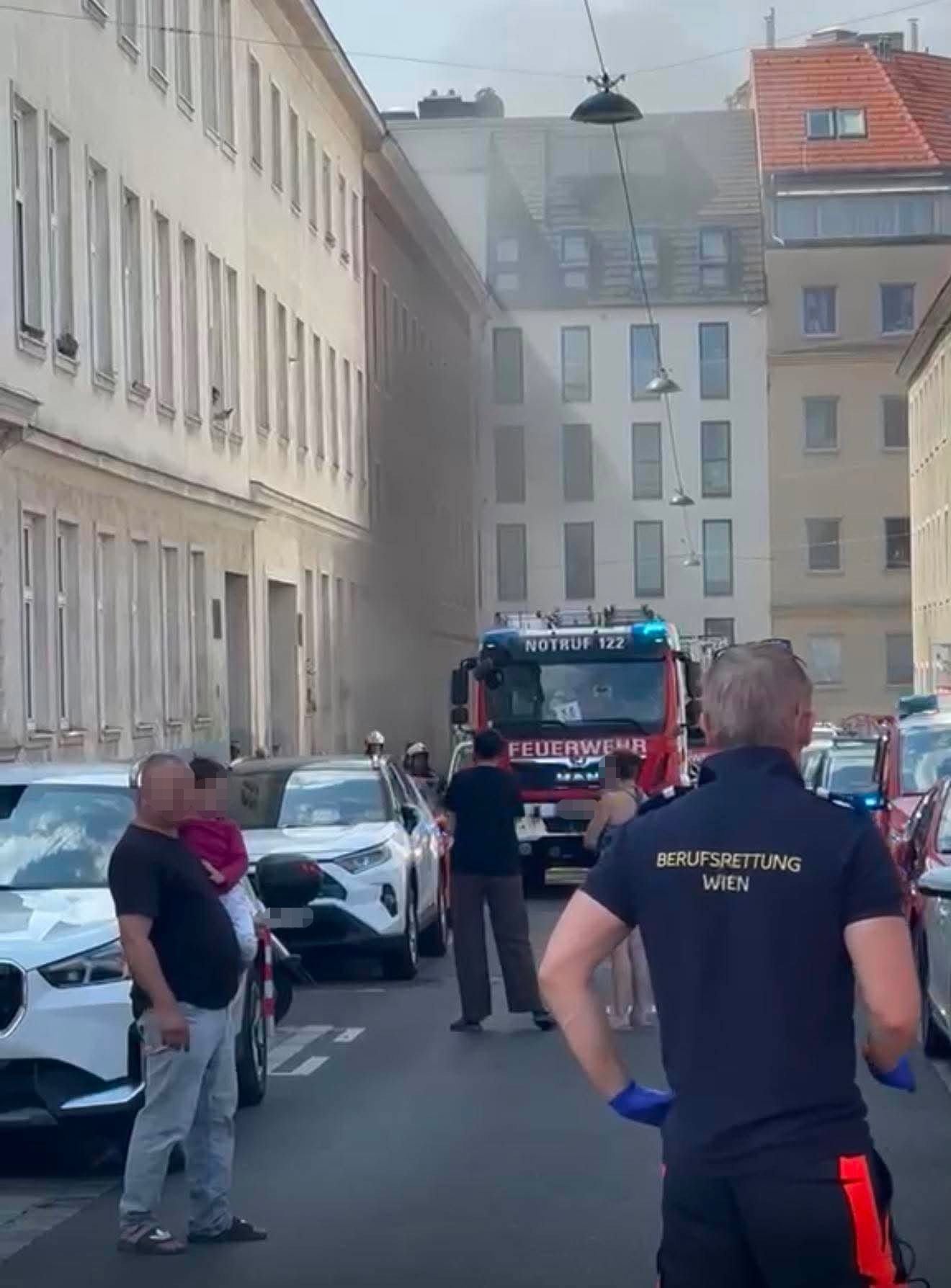Feuerwehreinsatz in einer verrauchten Straße, Menschen beobachten, Einsatzkraft von Wien im Vordergrund.