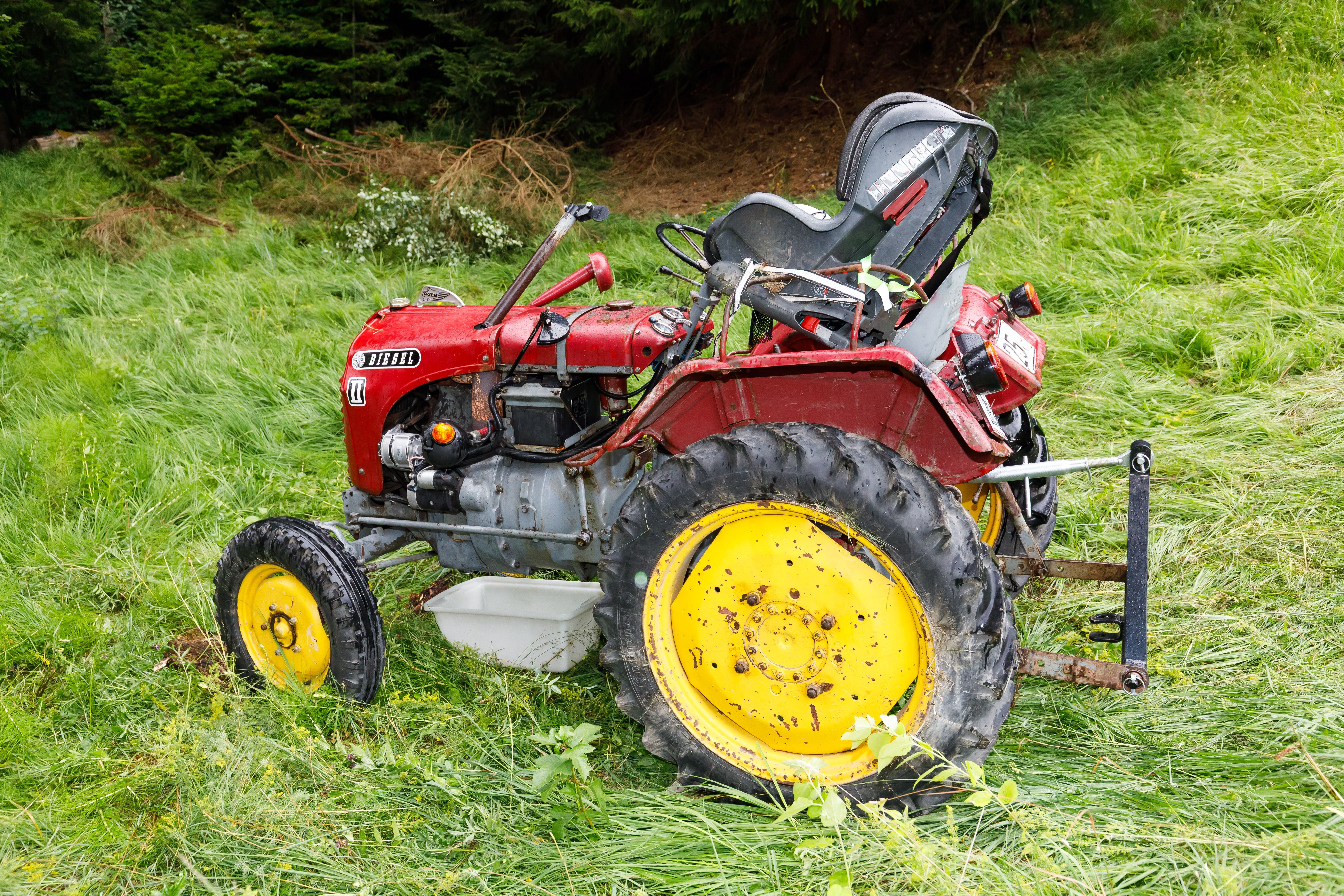 Roter Traktor mit gelben Rädern steht beschädigt auf einer Wiese in abschüssigem Gelände.