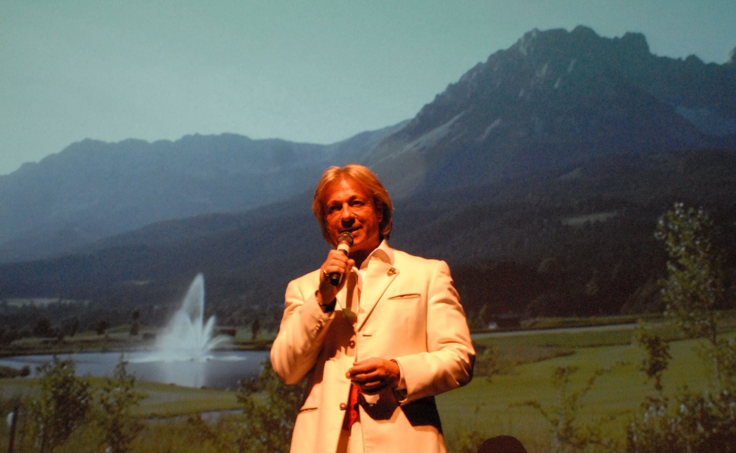 Mann im weißen Anzug singt auf Bühne mit Alpenlandschaft auf Videoleinwand im Hintergrund.