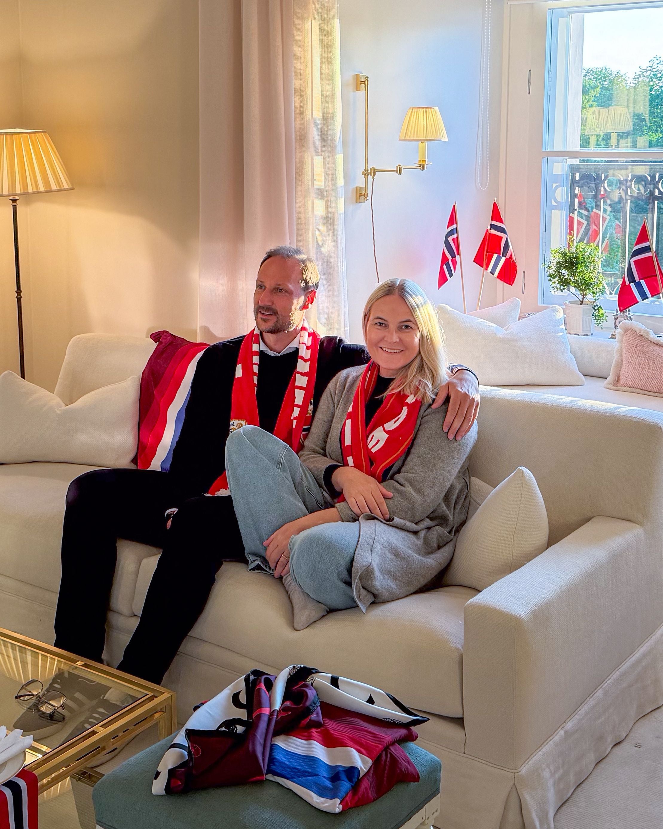 Ein Paar mit Norwegen-Schals sitzt auf einem Sofa und schaut ein Fußballspiel, norwegische Fahnen im Hintergrund.