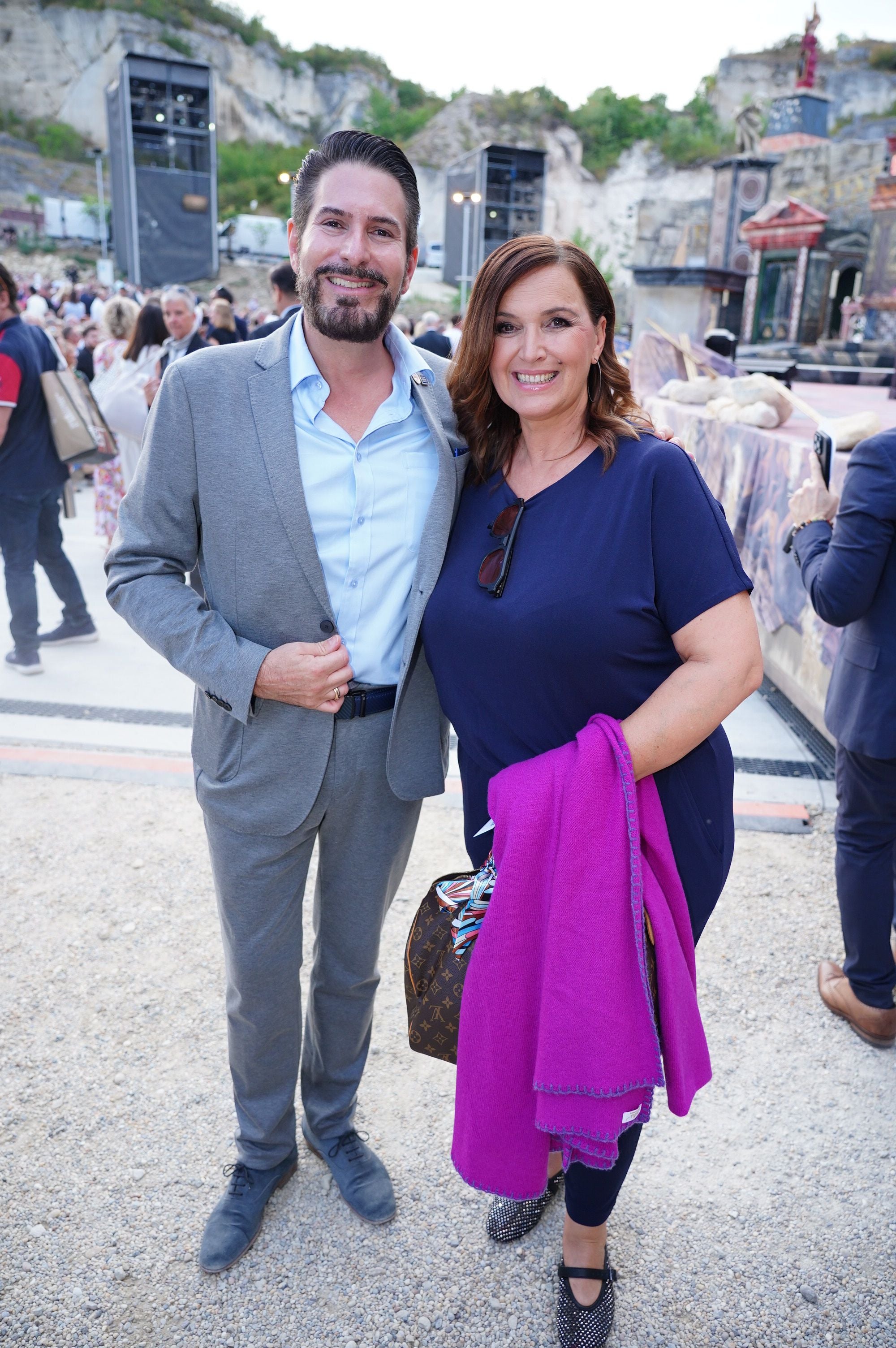 Ein Mann im grauen Anzug und eine Frau mit violettem Tuch lächeln bei einer Tosca-Premiere im Freien.