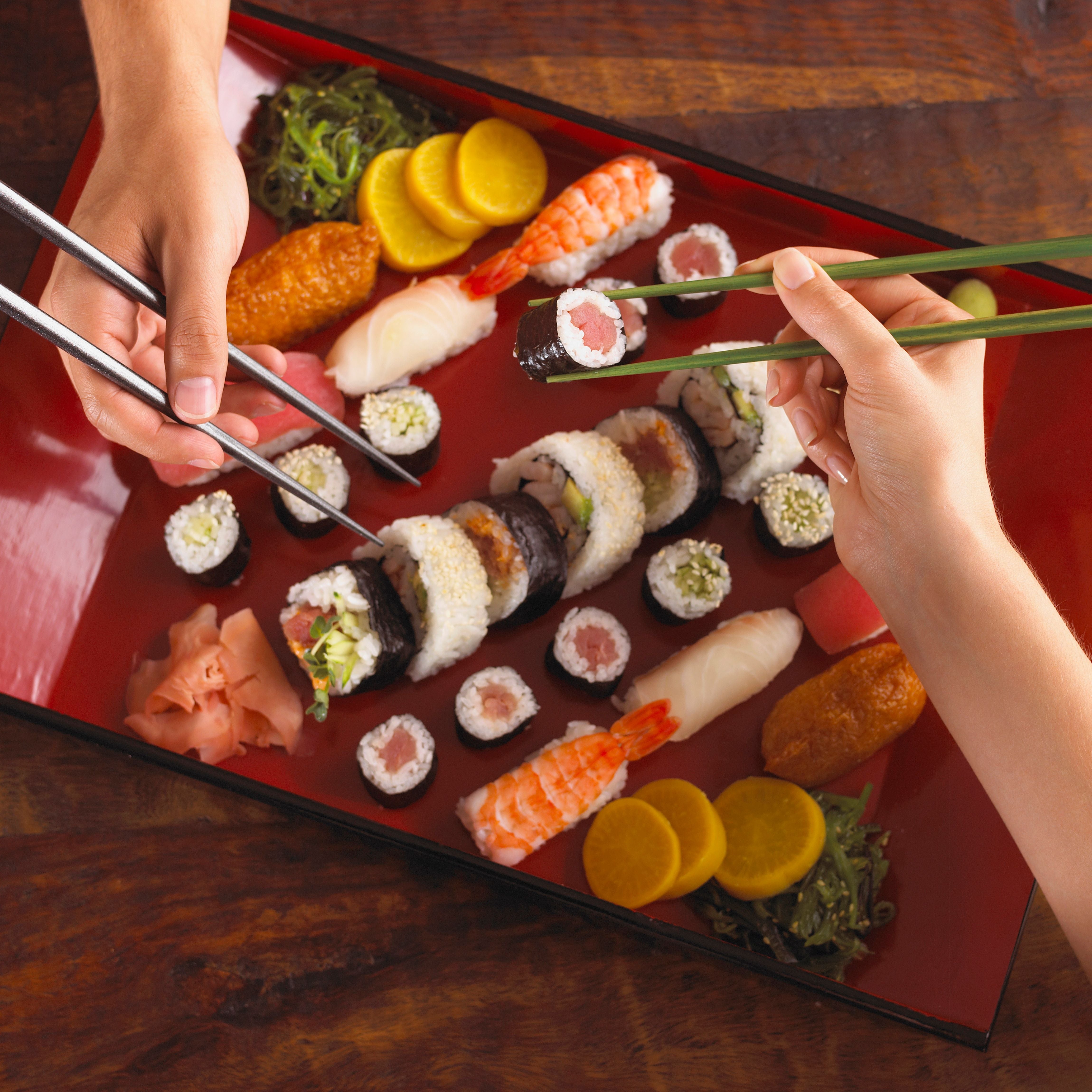 Zwei Hände nehmen Sushi mit Stäbchen von einer roten Platte voller verschiedener Sushi-Sorten.