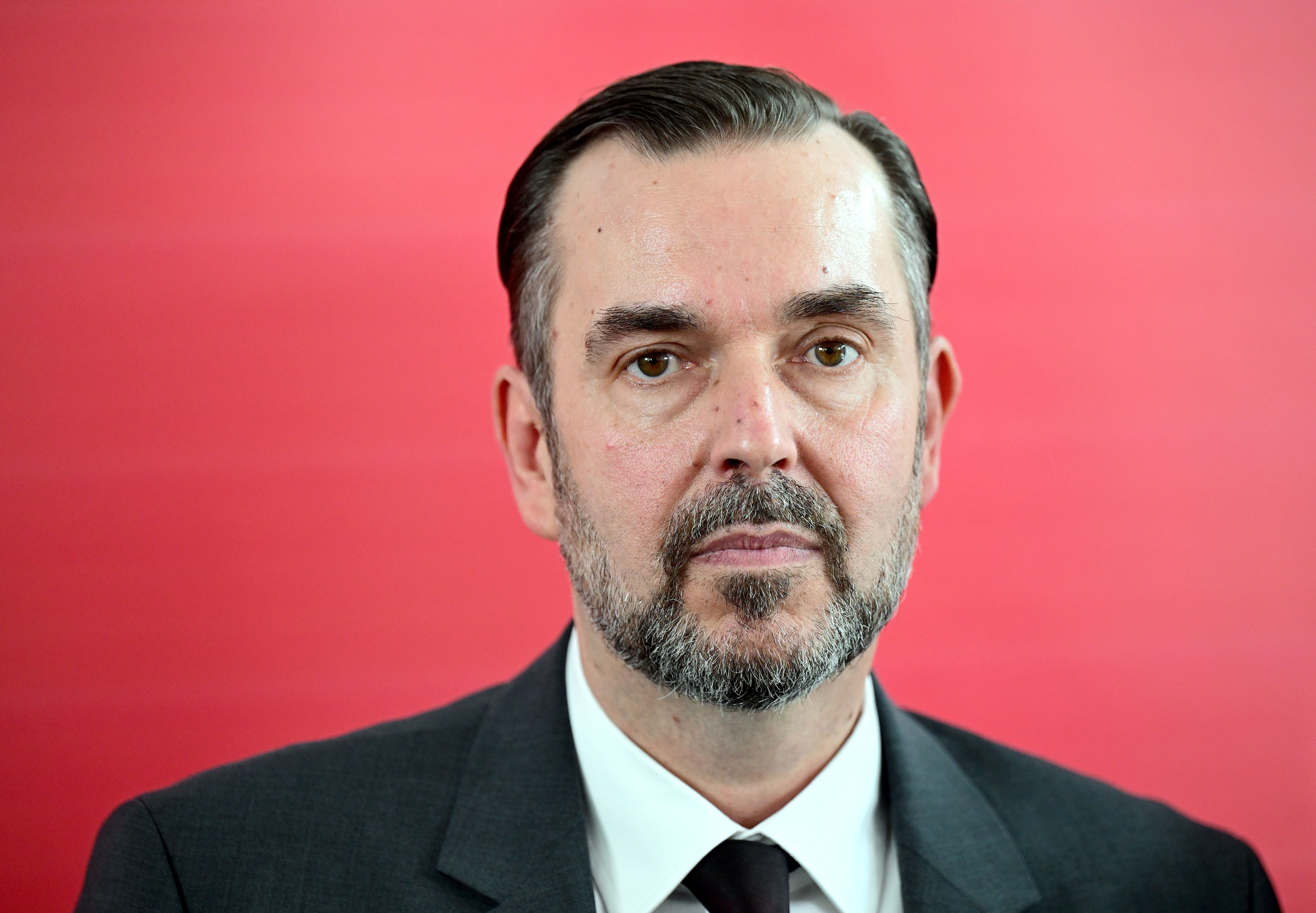 Ein Mann im Anzug vor rotem Hintergrund, neutraler Gesichtsausdruck.