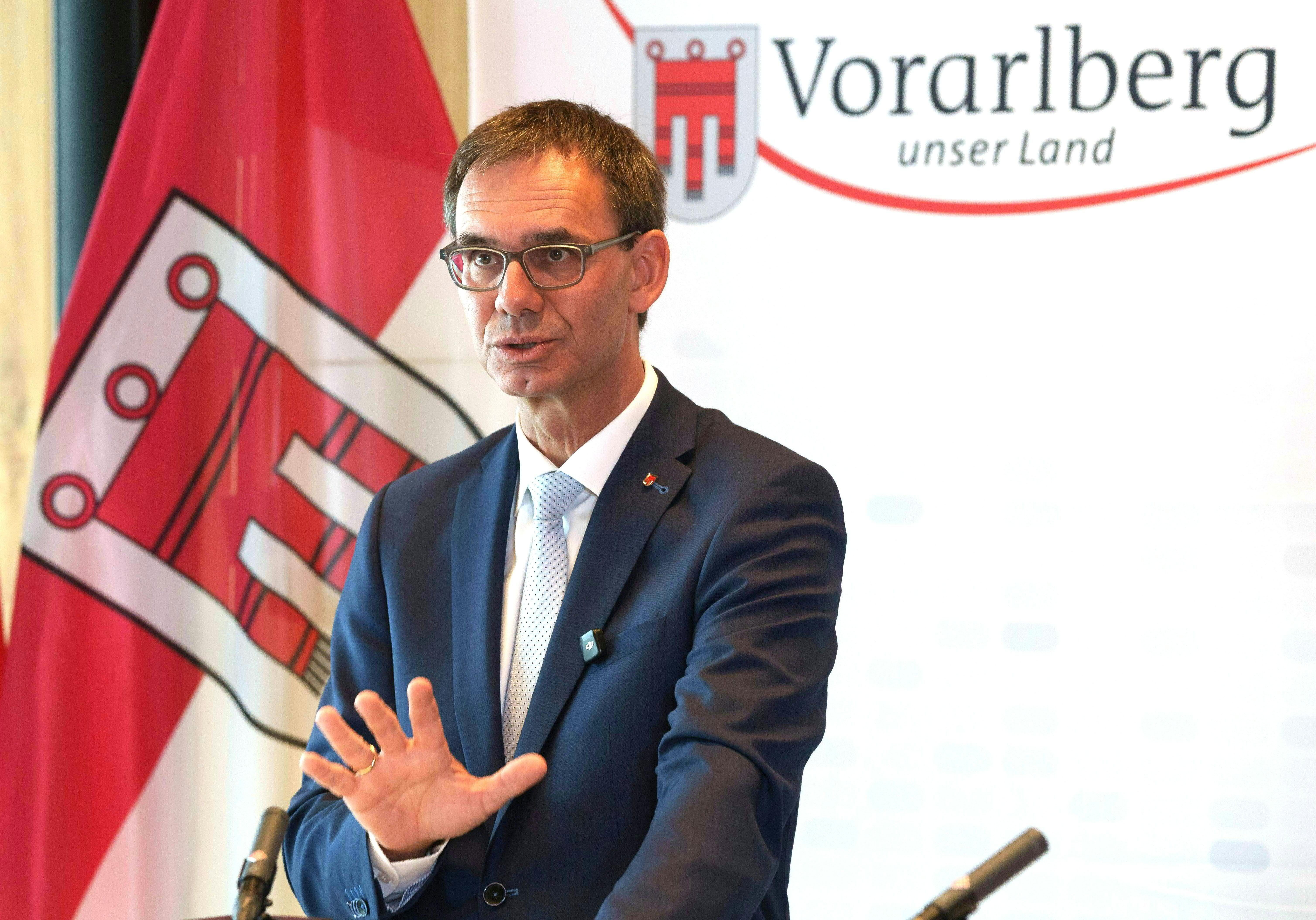 Vorarlbergs Landeshauptmann hält eine Pressekonferenz vor Logo und Flagge von Vorarlberg.