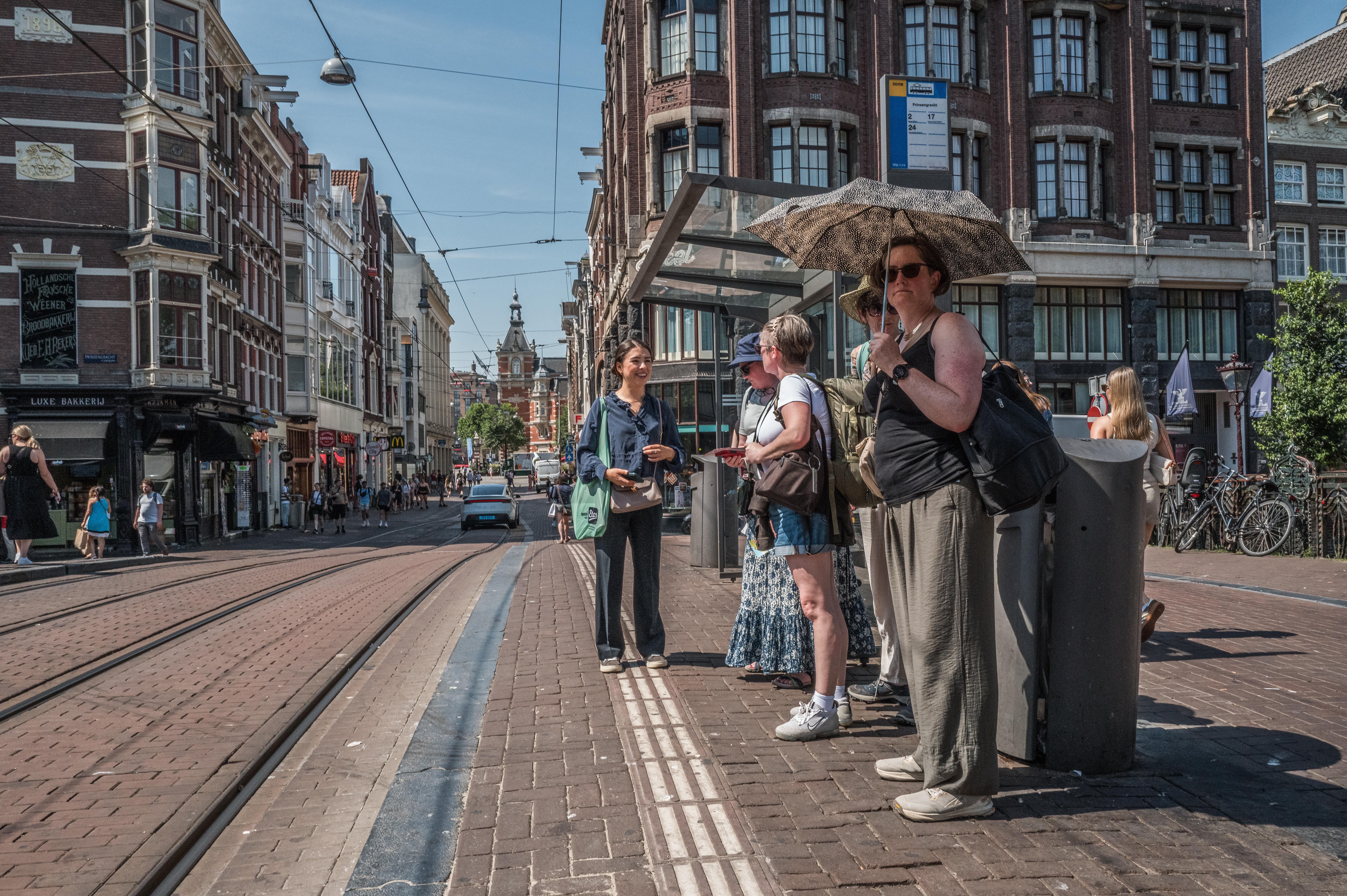 Touristen warten bei Hitze unter einem Sonnenschirm an einer Straßenbahnhaltestelle in Amsterdam.