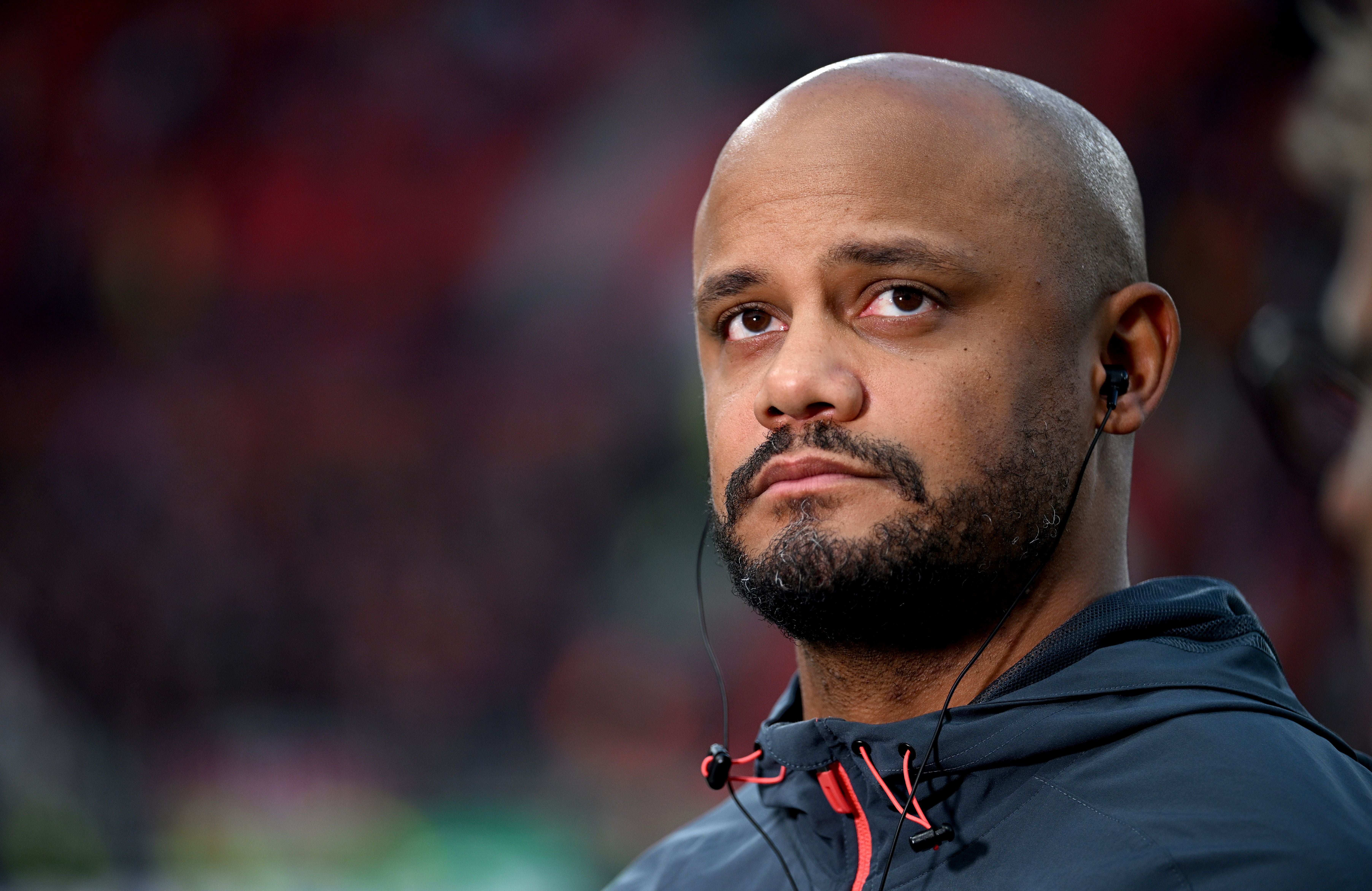 Ein Mann mit Bart und Kopfhörern blickt ernst, unscharfer Hintergrund im Stadion.