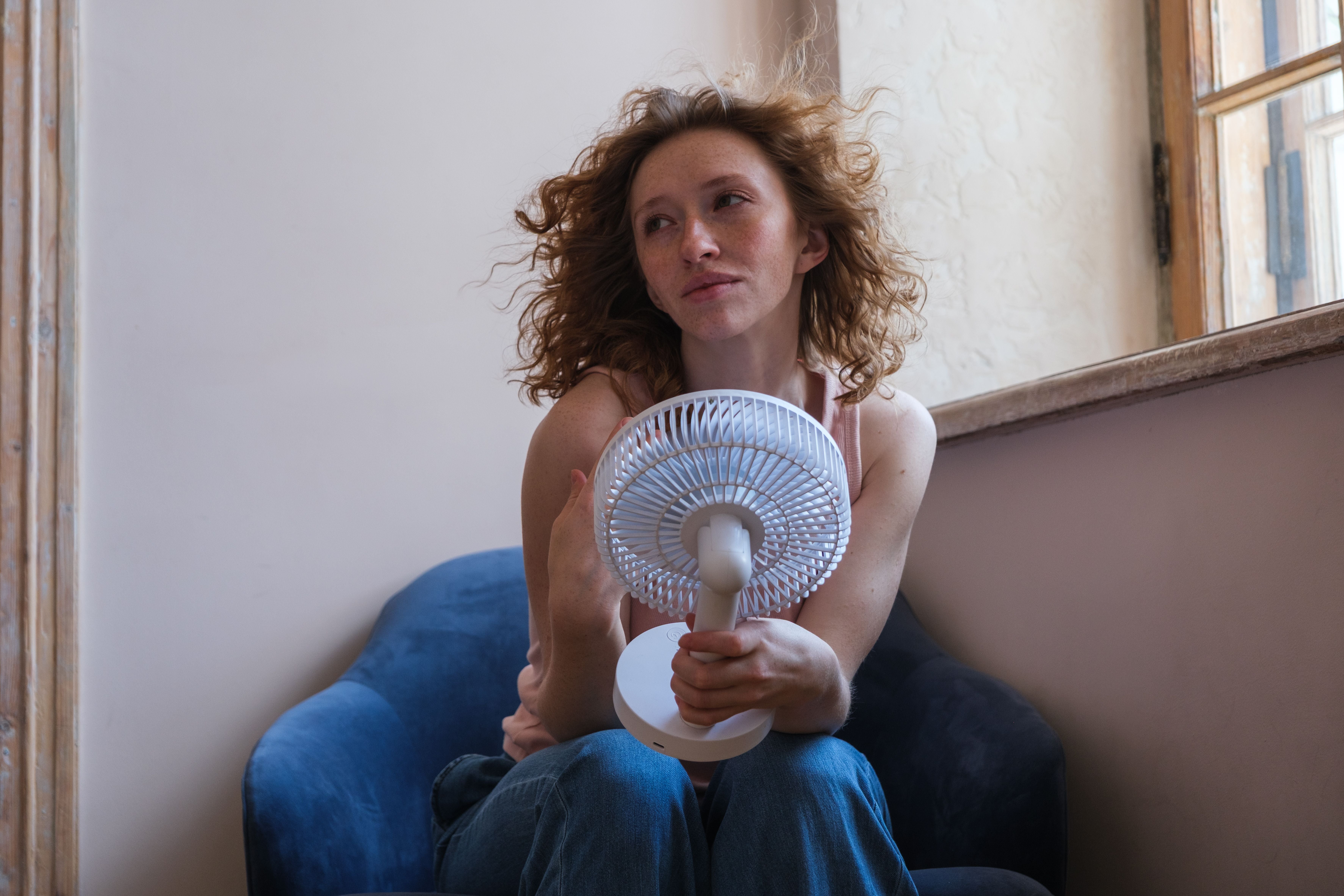 Frau mit roten Locken kühlt sich mit Ventilator in einem Raum ohne Klimaanlage ab.