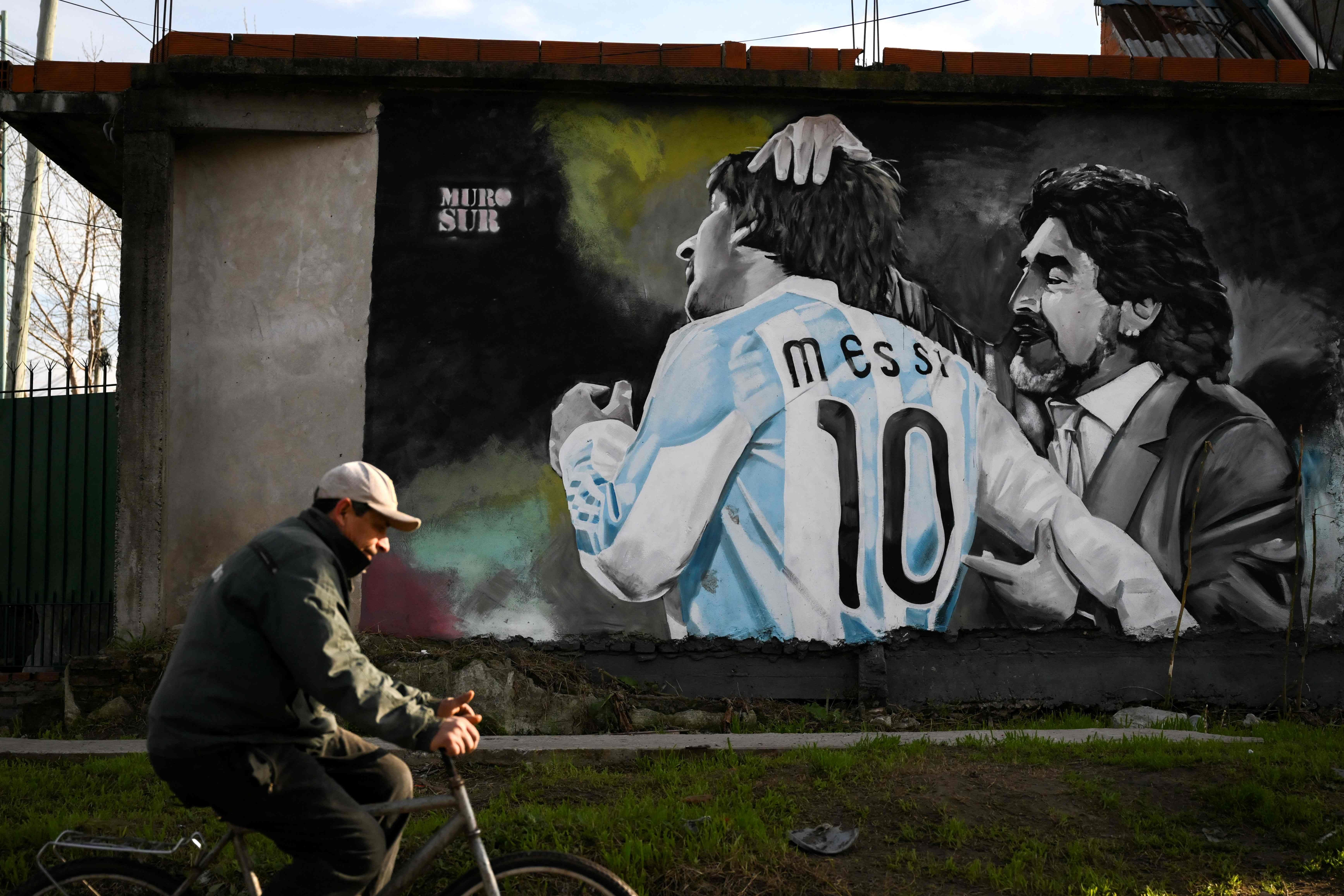 Ein Mann fährt Fahrrad an einem Wandbild von Maradona und Messi in argentinischen Trikots vorbei.