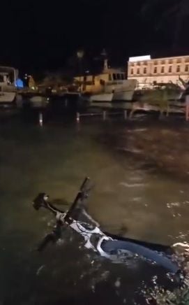 Ein Fahrrad liegt teilweise unter Wasser an einem Ufer bei Nacht, im Hintergrund Boote und Gebäude.