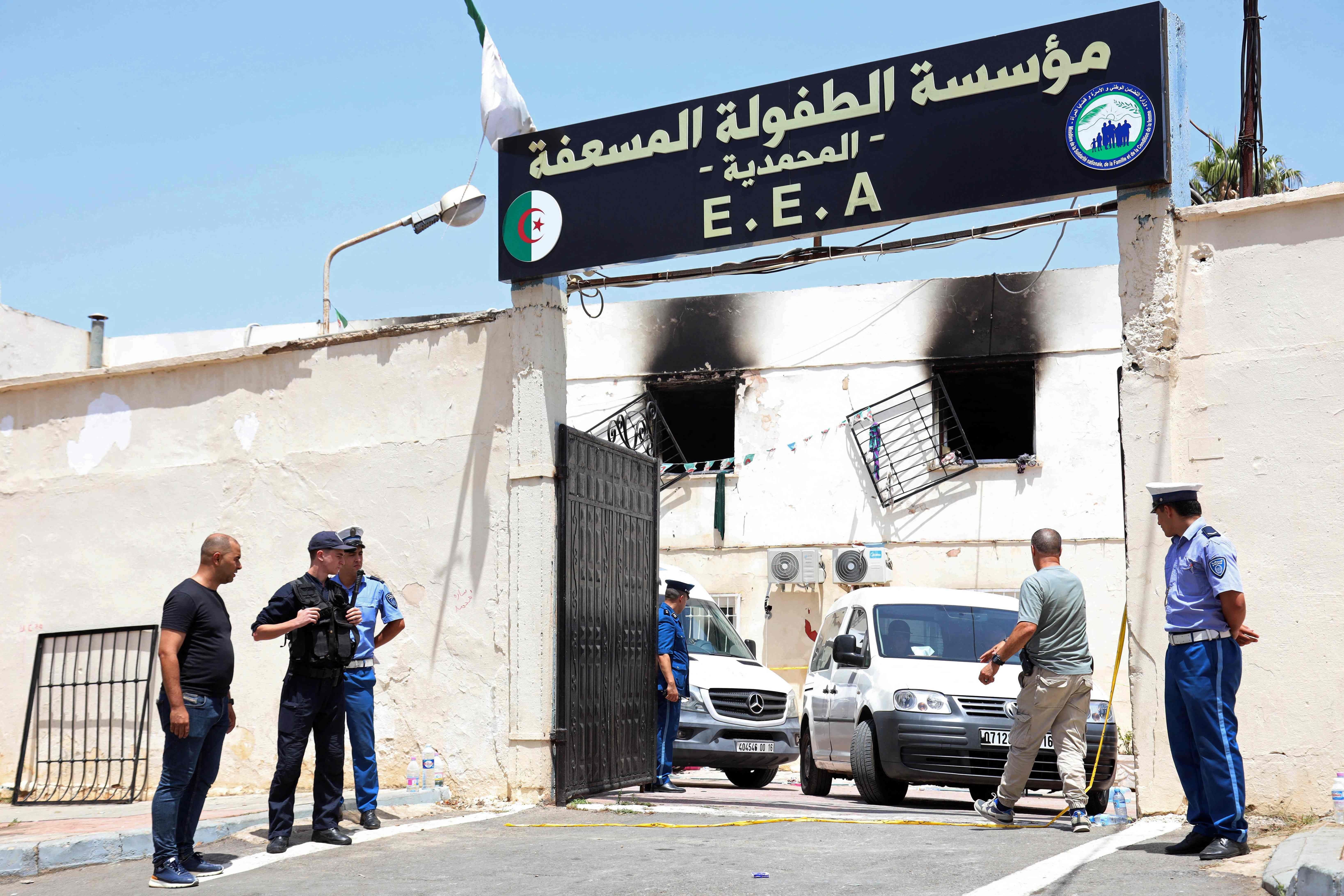 Polizisten und Männer stehen vor einem ausgebrannten Waisenhaus in Algier mit verkohlten Fenstern.