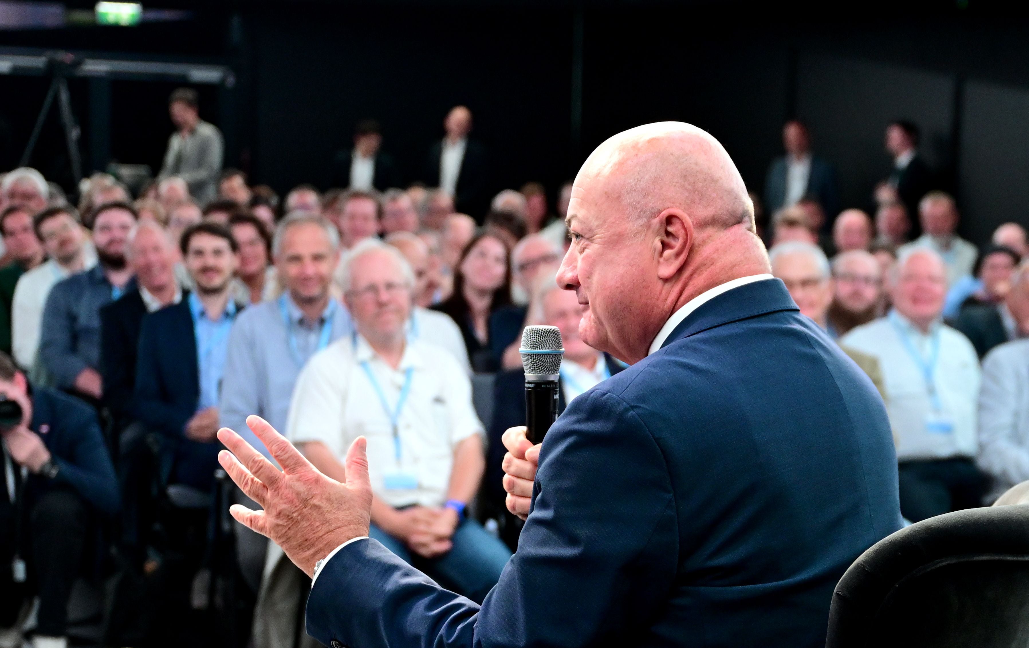 Ein Mann im Anzug spricht mit Mikrofon vor einem sitzenden Publikum bei einer Veranstaltung.