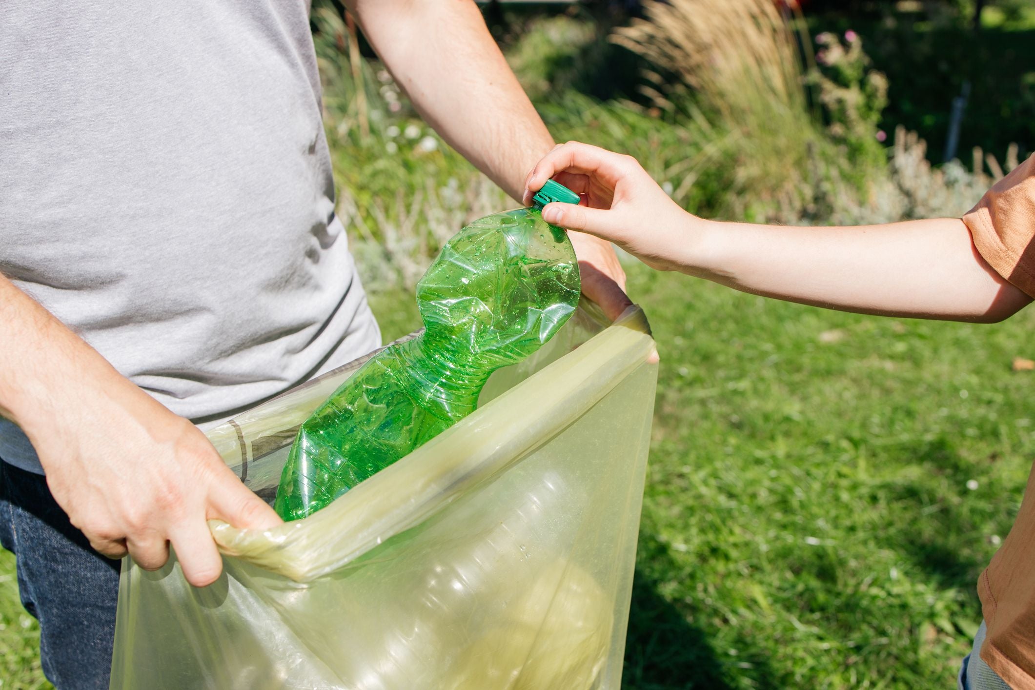 Hand wirft grüne Plastikflasche in einen gelben Müllsack im Freien.