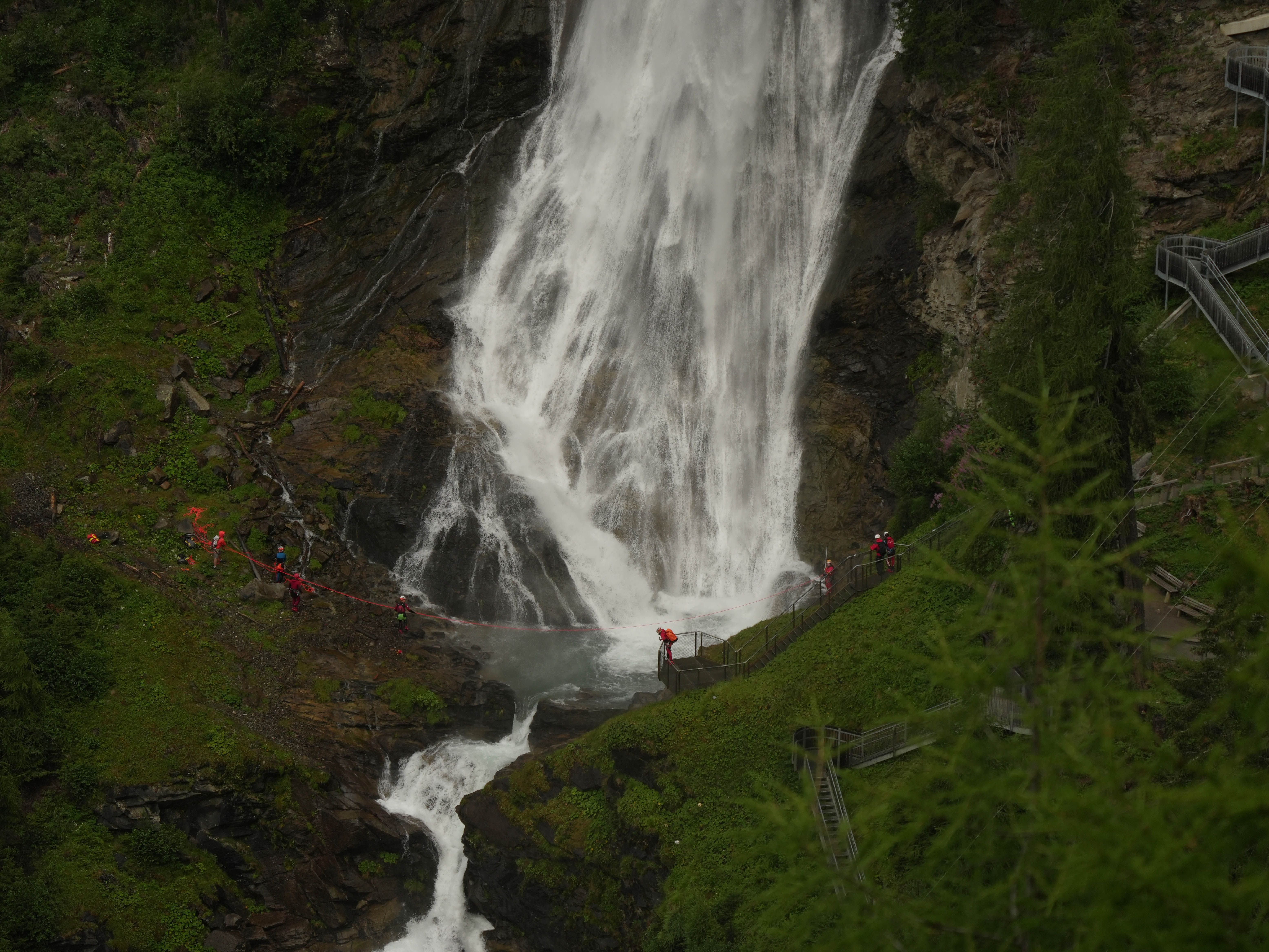 Menschen mit Sicherheitsausrüstung stehen bei einem großen Wasserfall in bergiger, grüner Landschaft.