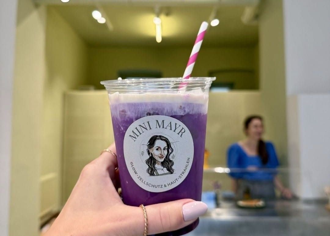 Eine Hand hält einen lila Ube-Latte mit Strohhalm und Mini Mayr-Logo im Vordergrund.