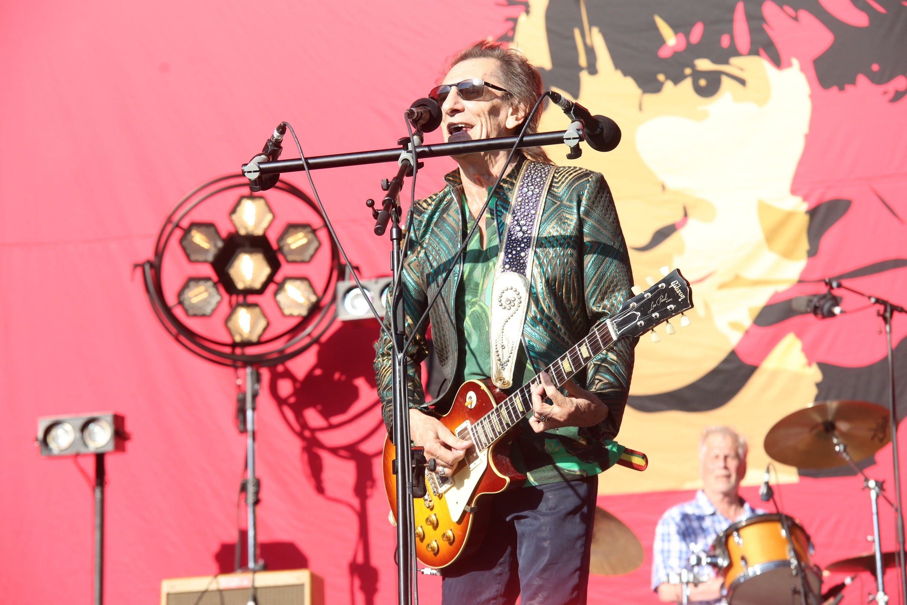 Ronnie Wood spielt E-Gitarre und singt auf einer Bühne mit rotem Hintergrund und Schlagzeuger.