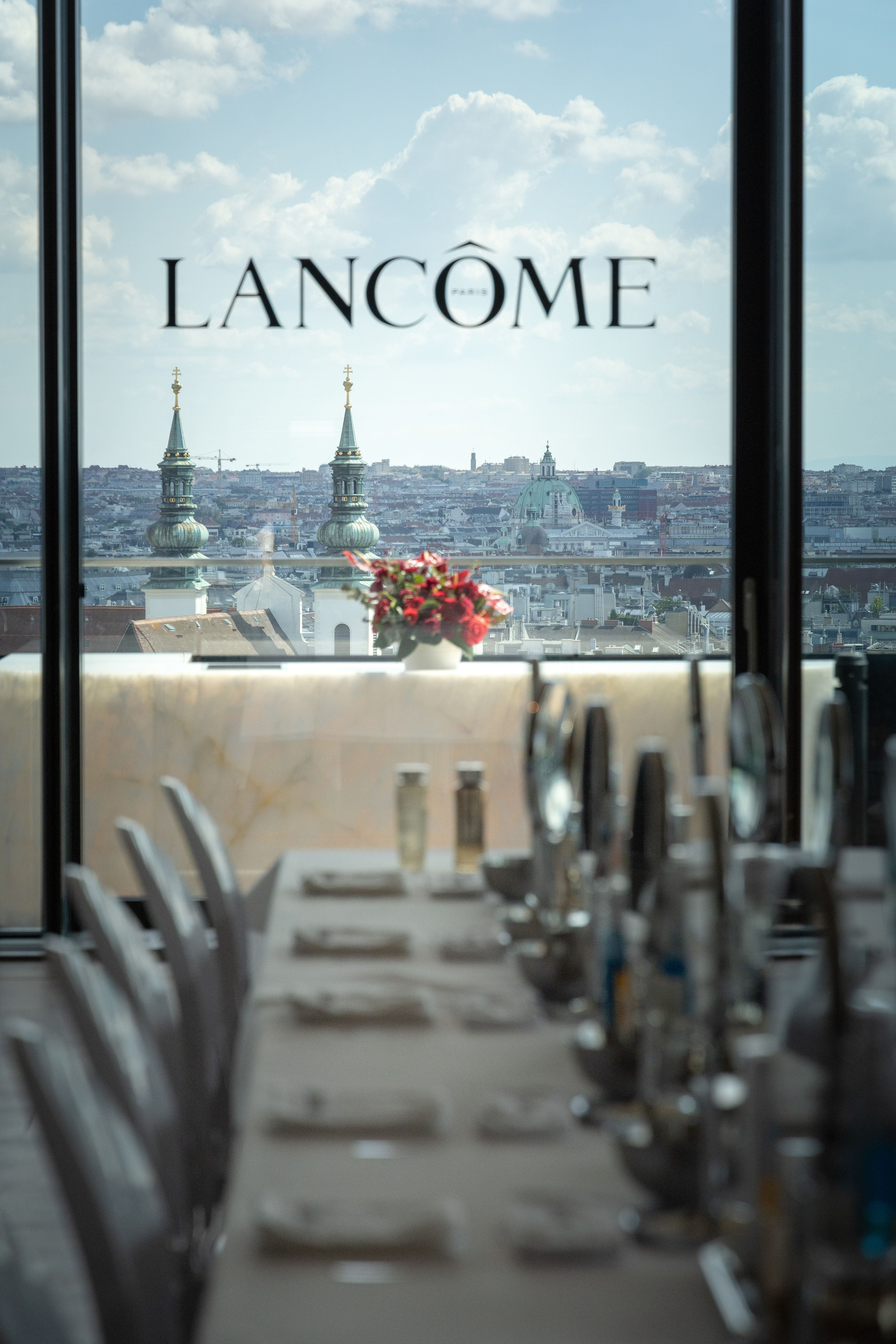 Elegant gedeckter Tisch vor Fenster mit Lancome-Logo und Blick über eine Stadt mit Kirchtürmen.