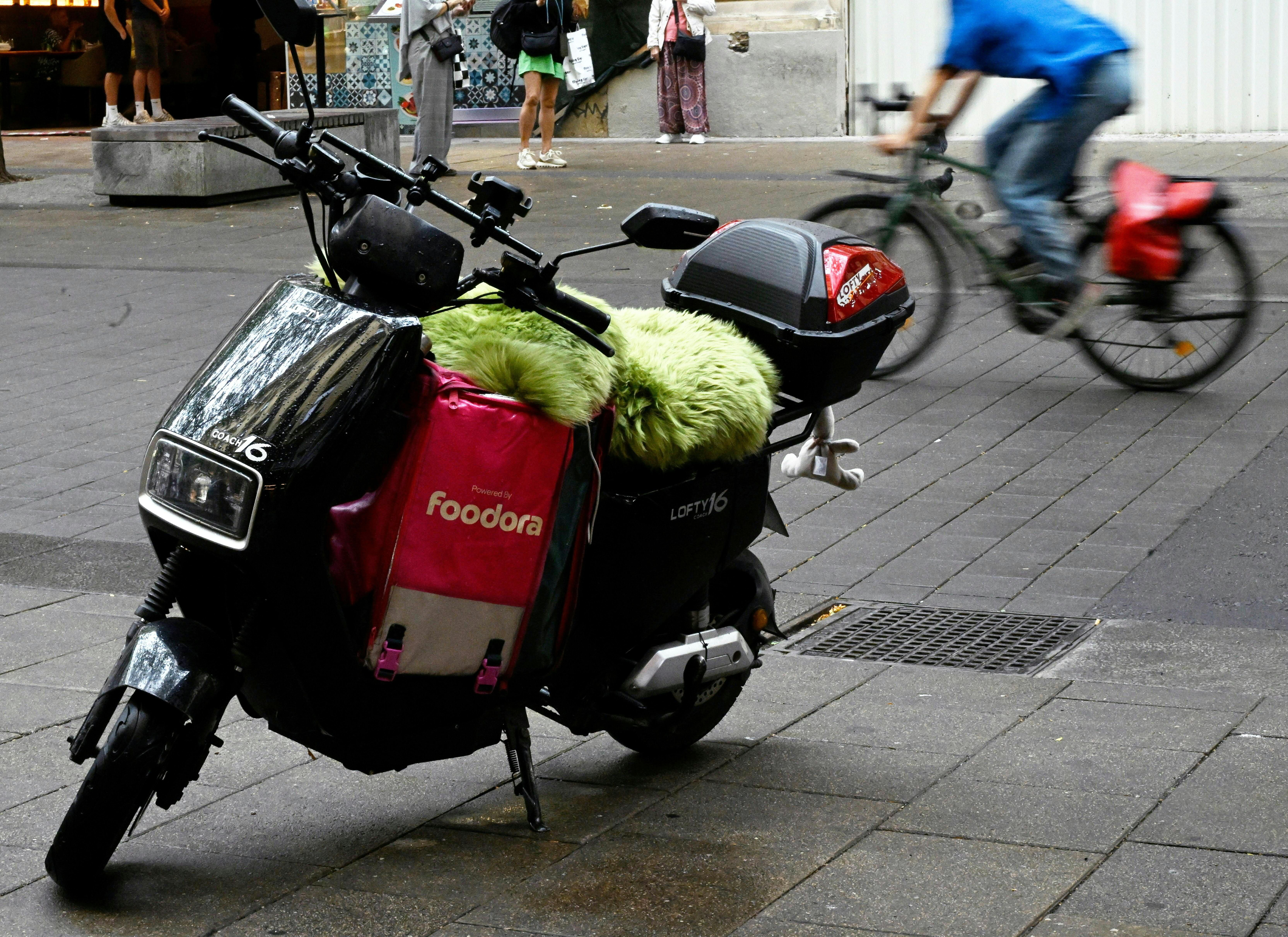Ein E-Moped mit Foodora-Liefertasche steht auf einer Straße in Wien, im Hintergrund ein Radfahrer.