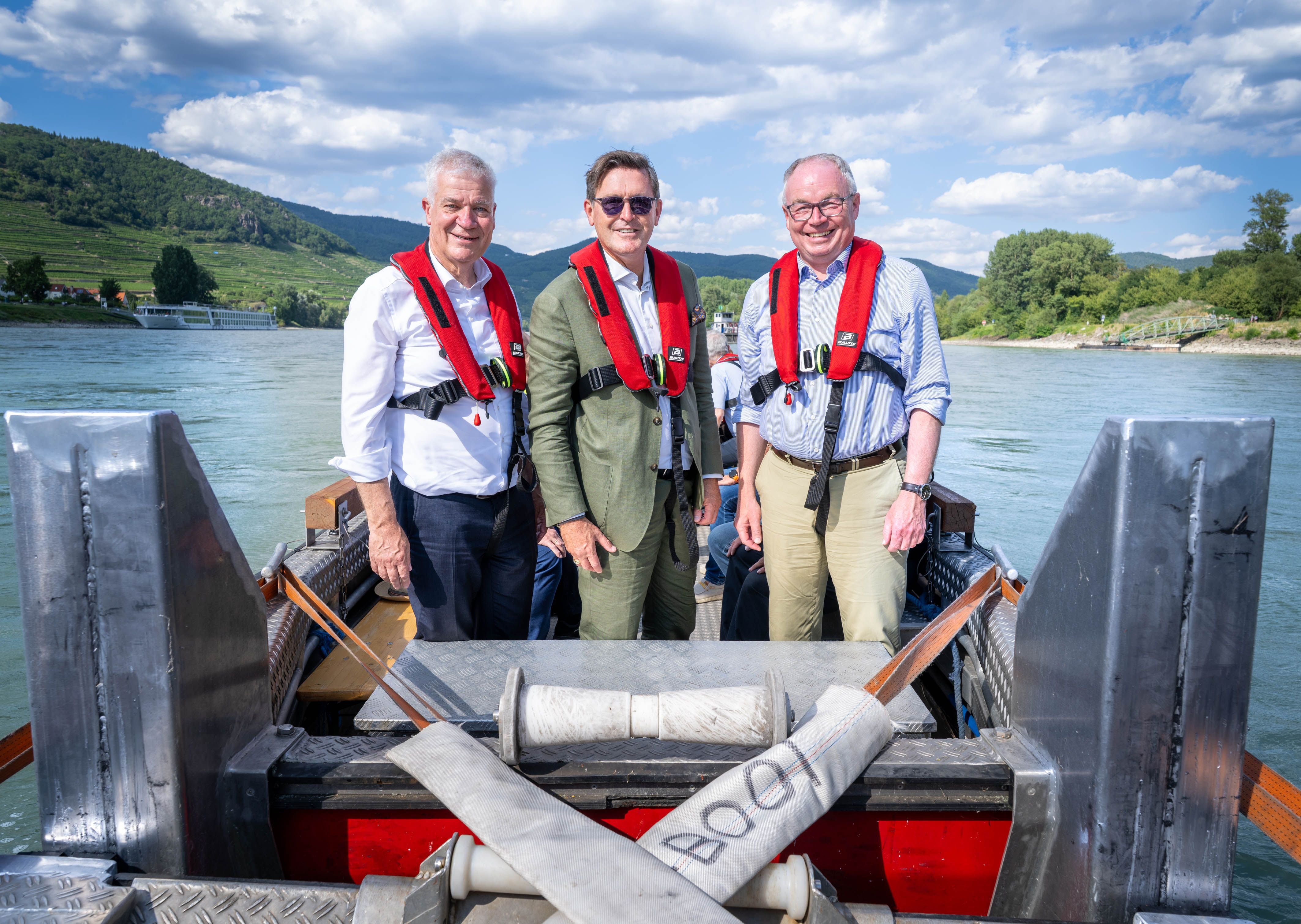 Drei Männer mit roten Rettungswesten stehen auf einem Boot auf einem Fluss vor bewaldeten Hügeln.